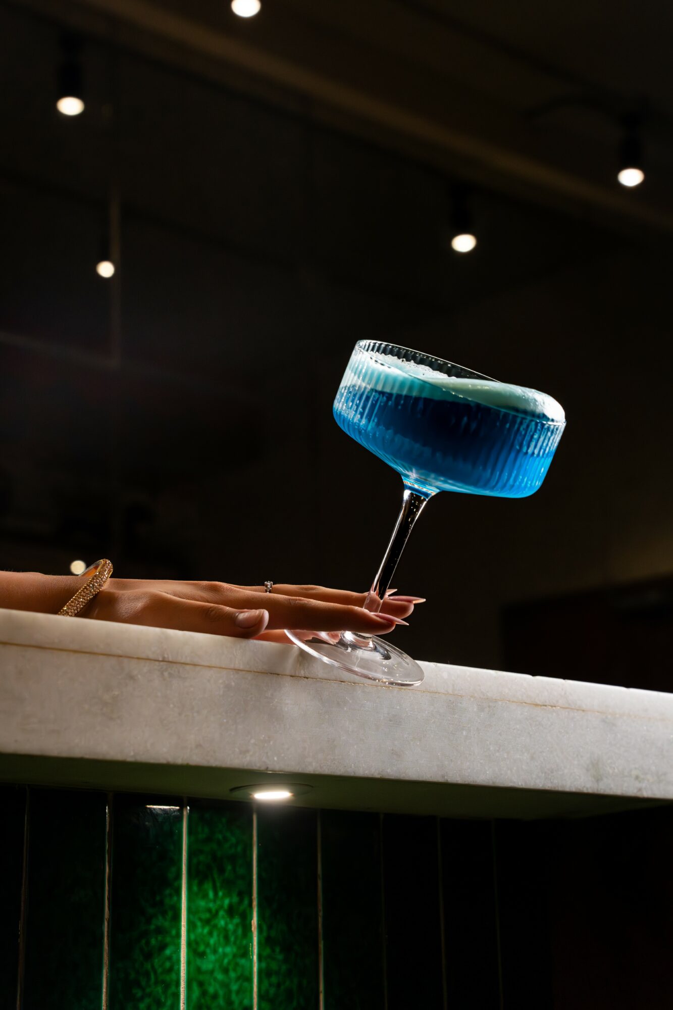 A hand with red nail polish holds a blue cocktail in a coupe glass on a white surface, with a dark background and ceiling lights.