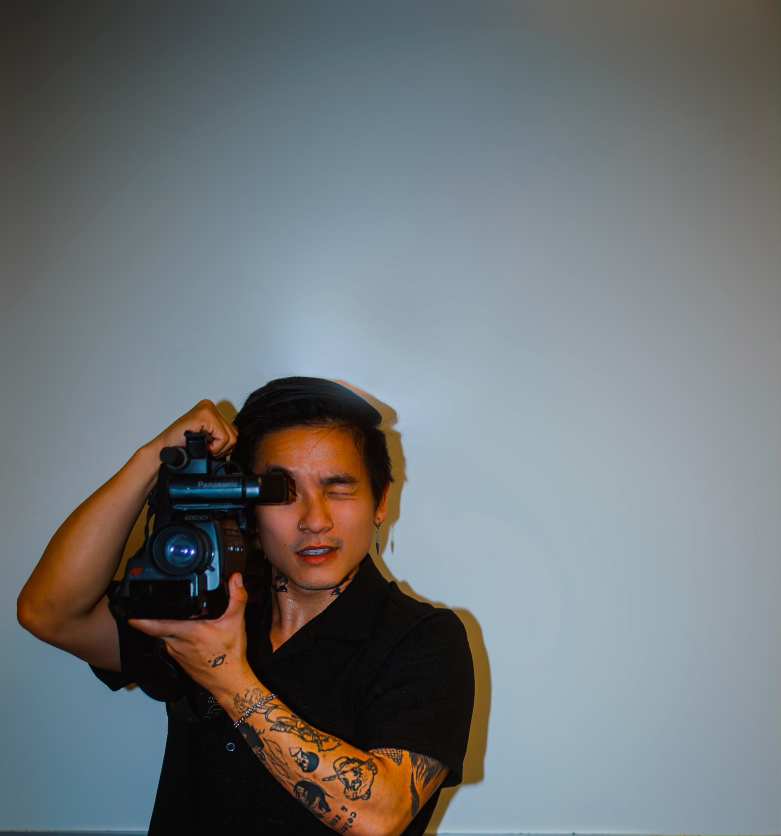 Person holding a camera, with tattoos on their arm, standing against a plain background.
