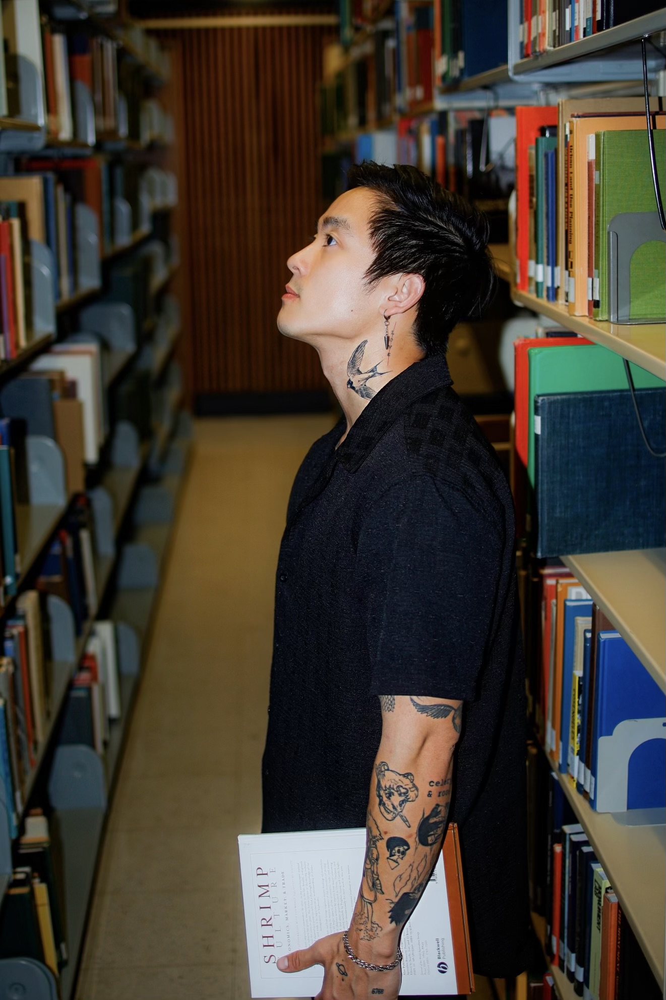 Person with tattoos on arm and neck, standing in a library aisle, looking upward, surrounded by bookshelves.