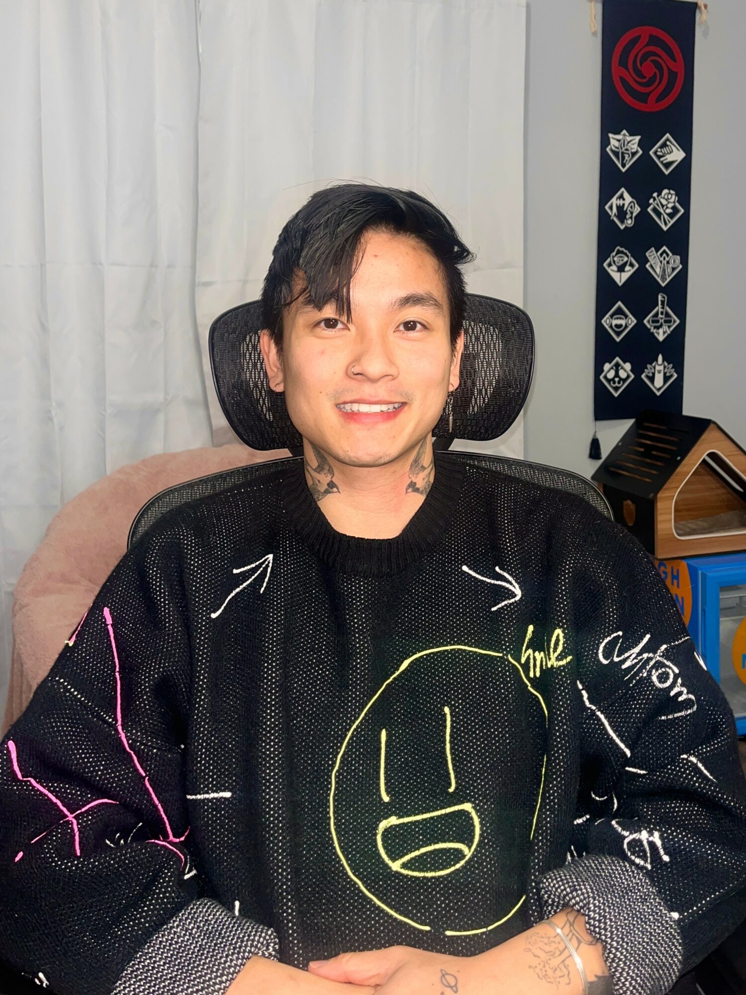 Young man with black hair smiling, sitting in a chair, wearing a black sweater with a yellow smiley face, in a room with curtains and a poster.
