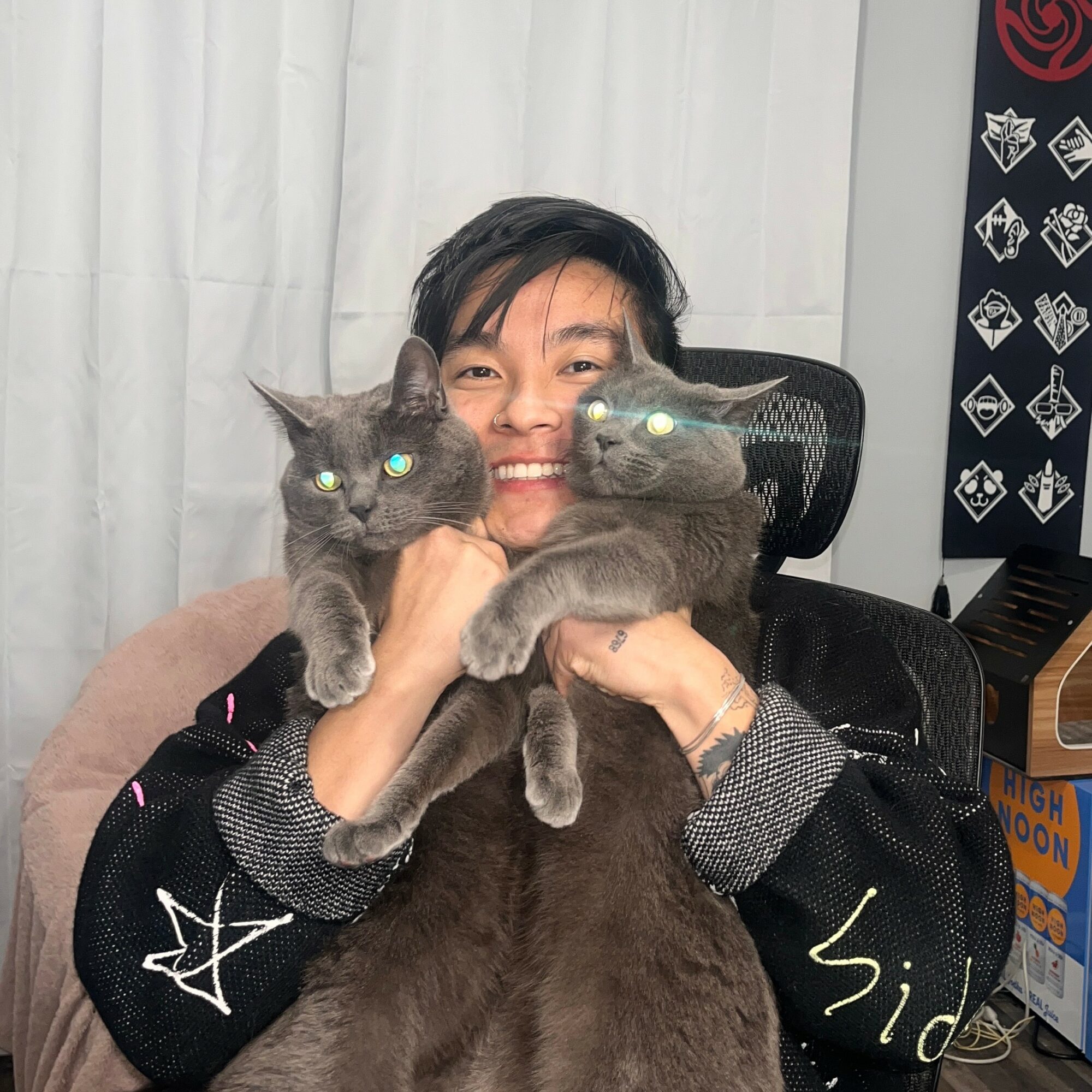 Person holding two gray cats indoors, smiling, with a black chair and patterned wall in background.