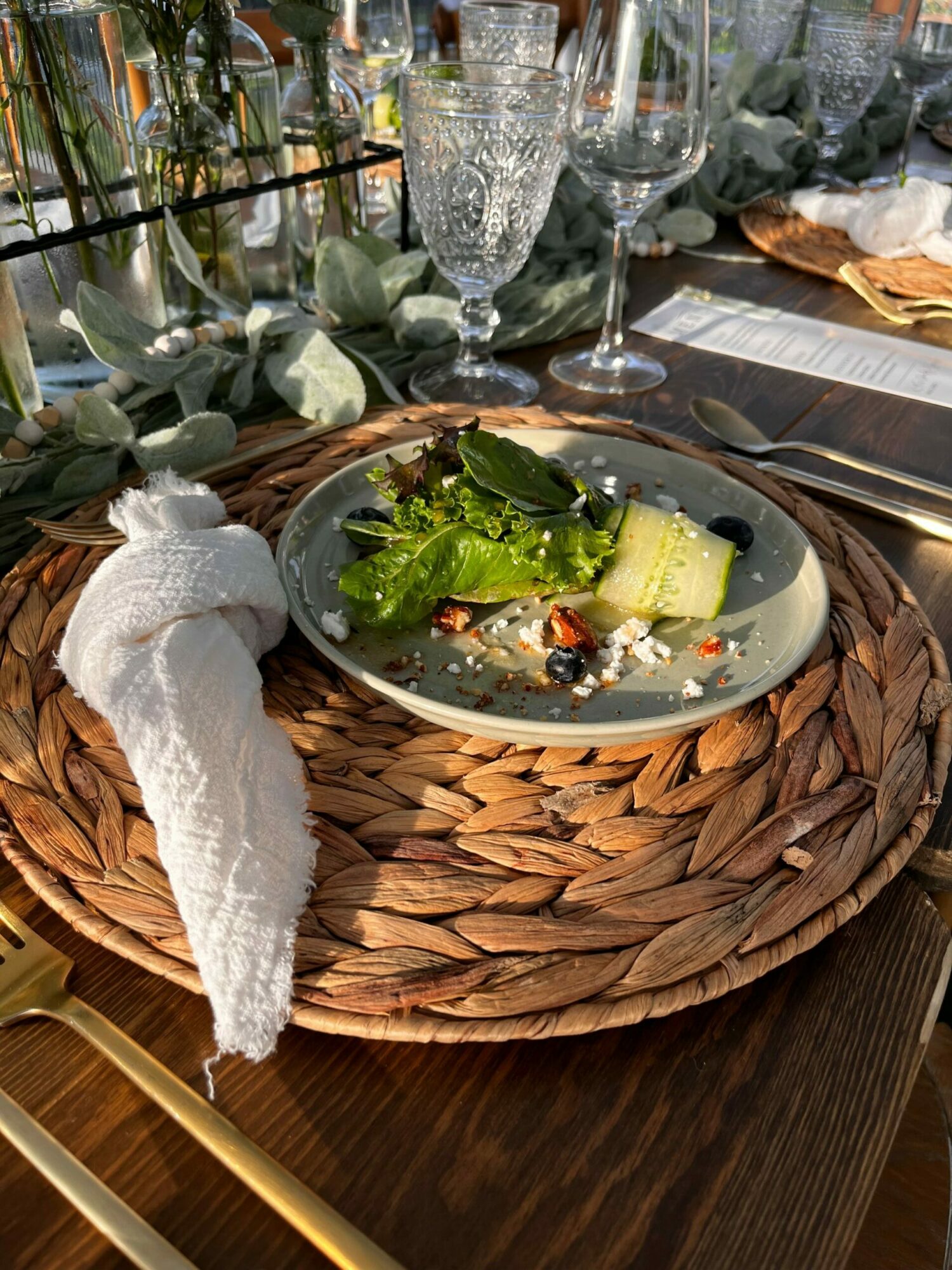 Empty plate with a cucumber slice, greens, and berries on a woven placemat, with a white napkin and glassware nearby.