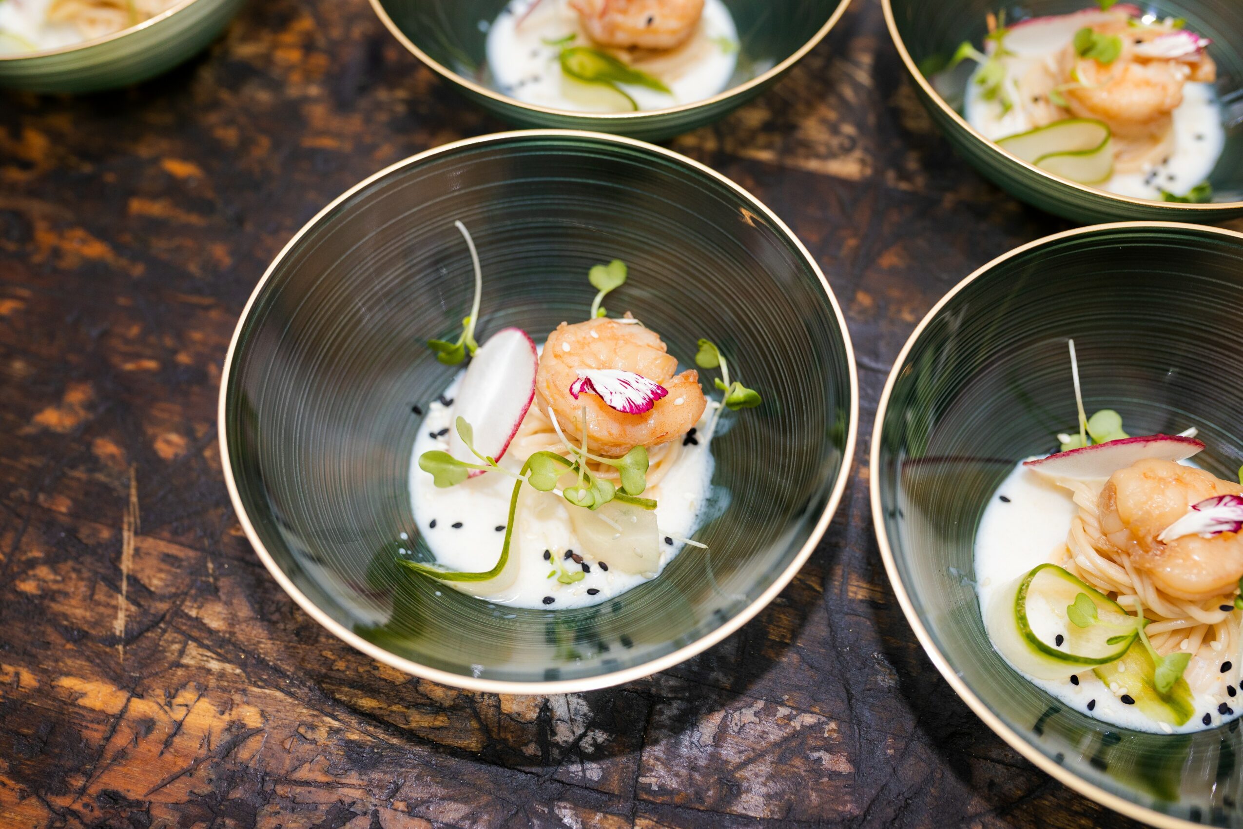 Four black bowls with food, garnished with pink, white, and green ingredients, on a dark textured surface.
