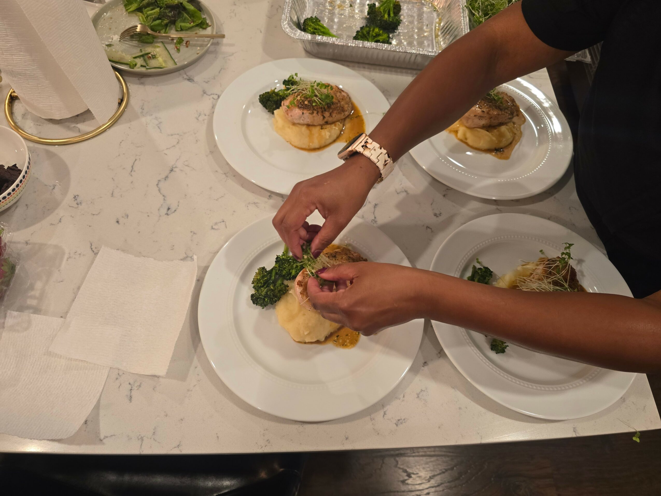Person arranging food on a white plate with other plates on a marble table, hands visible, wearing a watch.