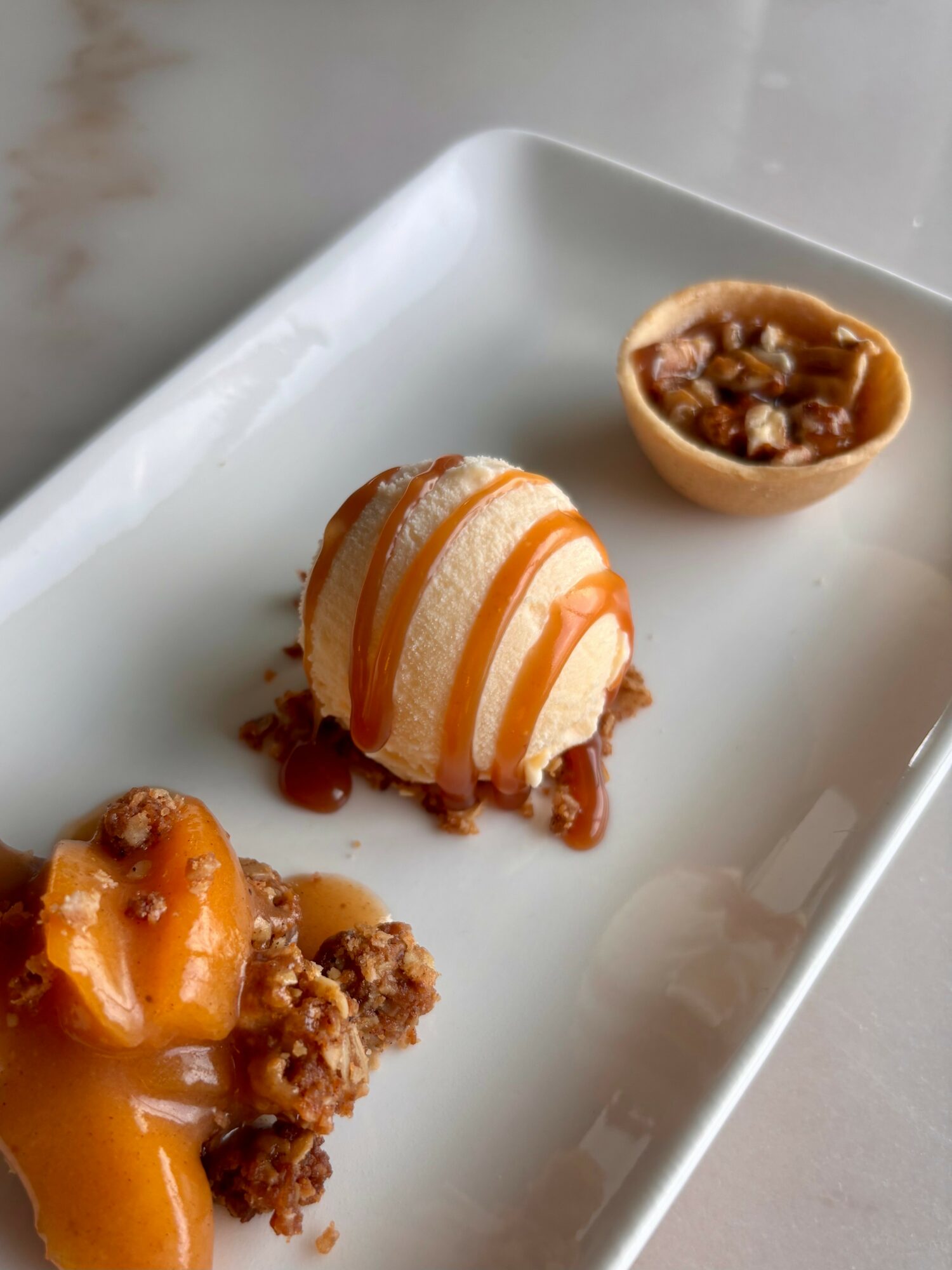 A dessert with a scoop of ice cream topped with caramel sauce, a small tart with caramel filling, and a crumbly topping on a white rectangular plate.