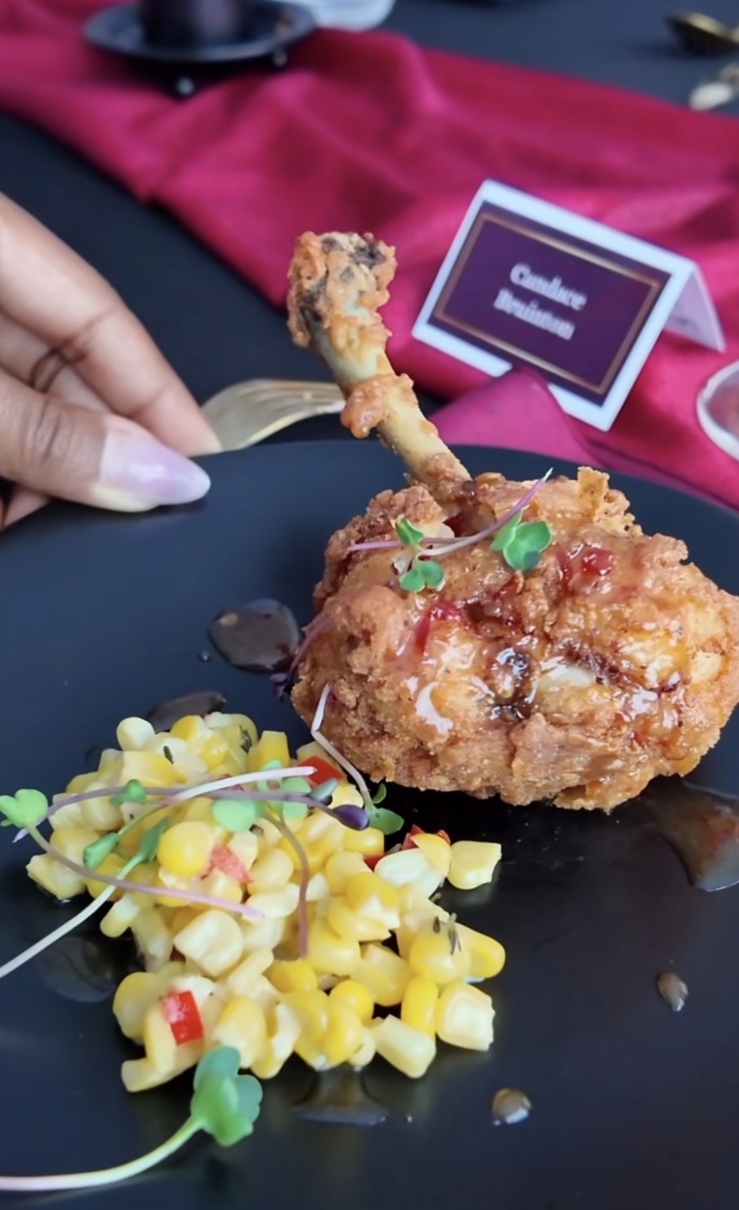 Hand holding a fried chicken drumstick with colorful candy decorations on a black plate, pink cloth and a small sign in the background.
