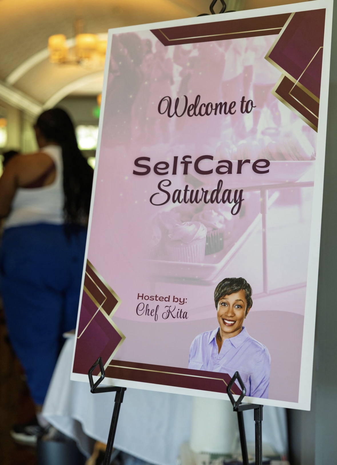 Event sign with a smiling child, welcoming to SelfCare Saturday, hosted by Chef Kita, with pink and maroon design elements.