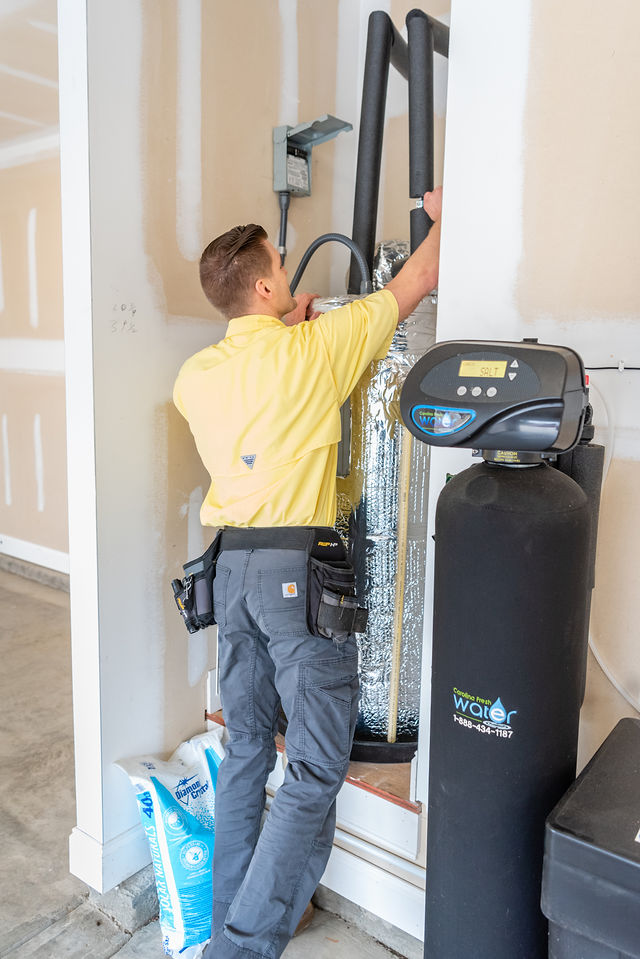 A man in a yellow shirt and gray pants operates a water softener system in a utility room.