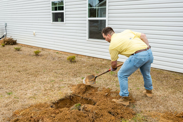 Man in yellow shirt and jeans digging a hole in a yard near a house with white siding and windows.
