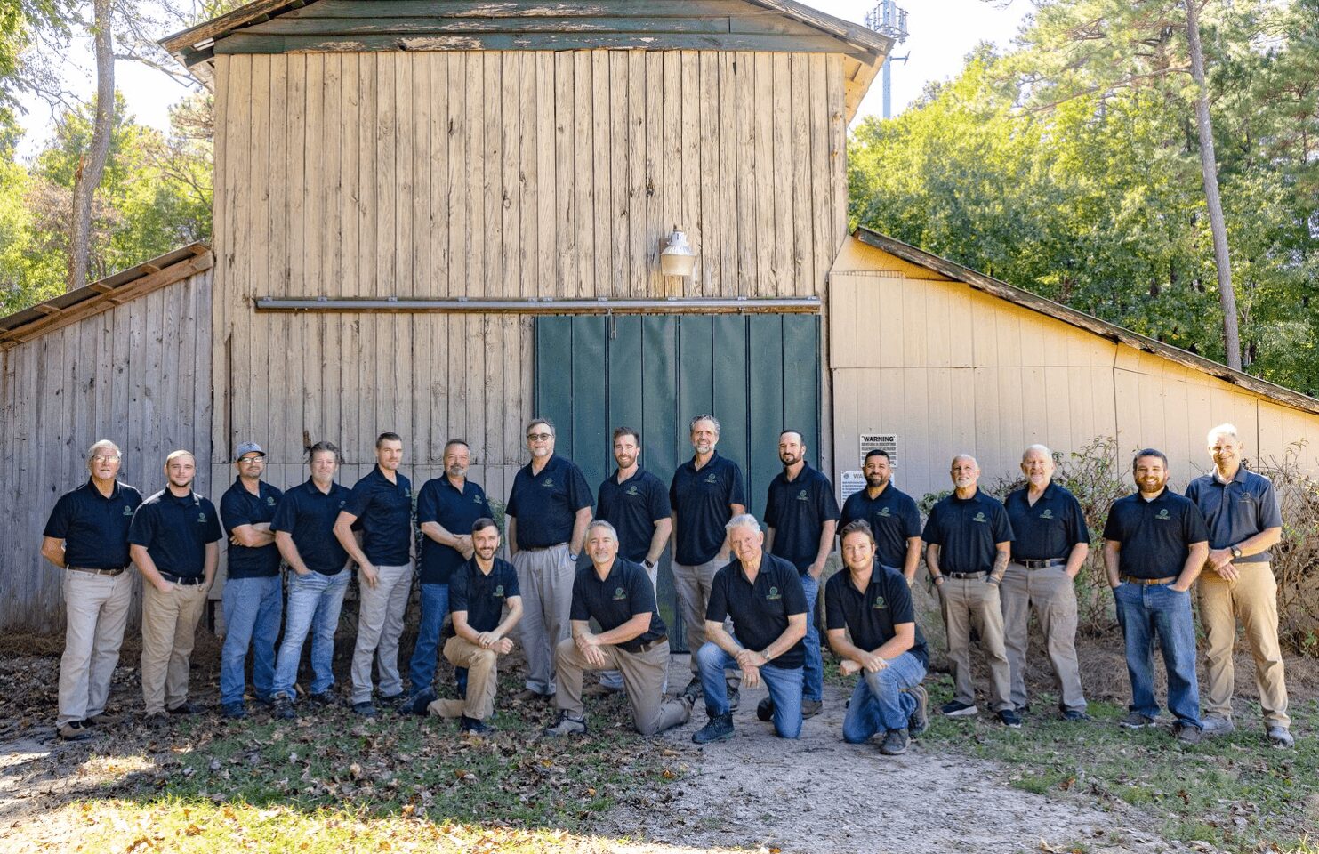 Group of 16 people standing in front of a large wooden barn, some with hands in pockets, outdoors on grass.