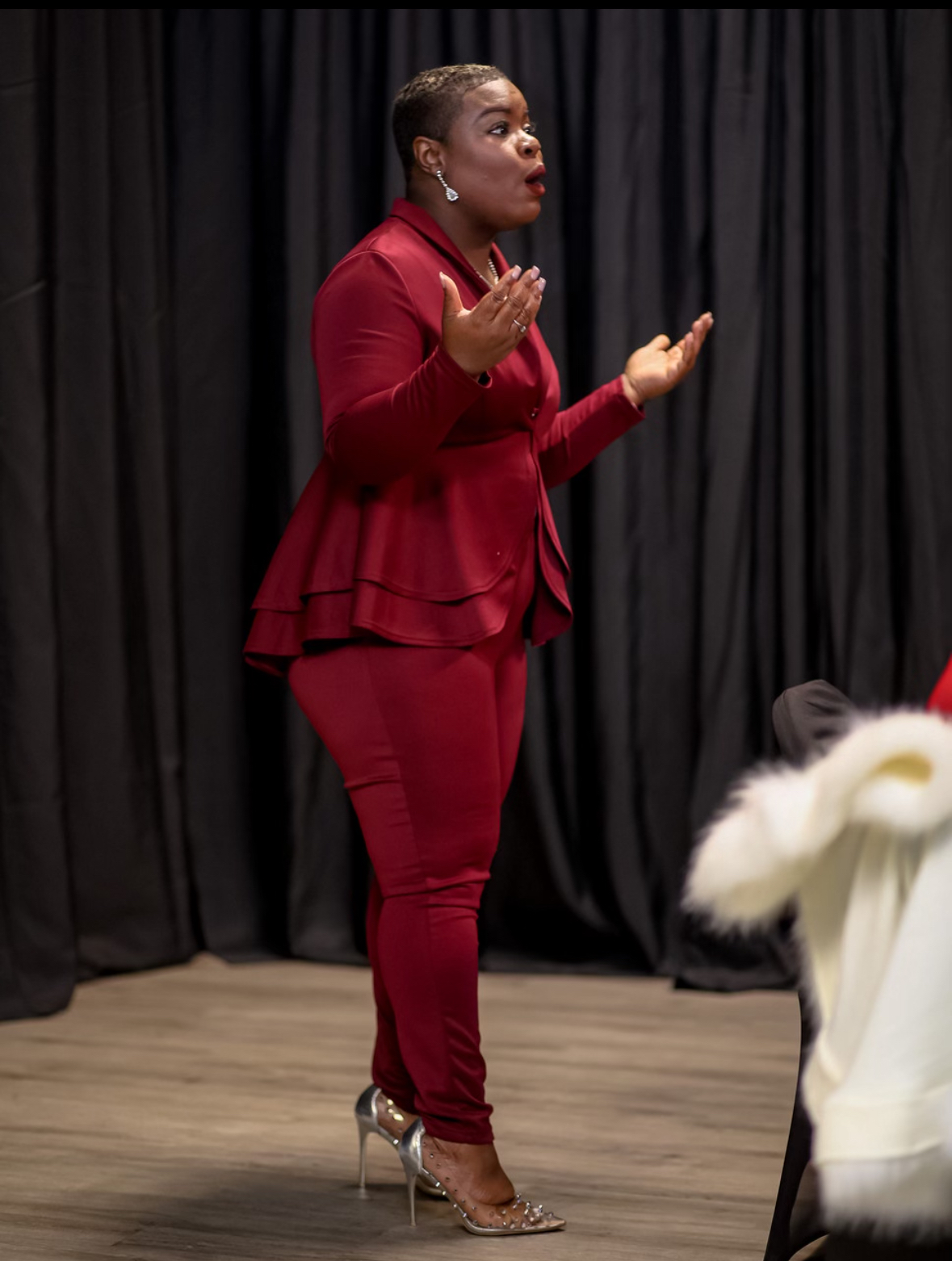 Woman in red outfit speaking with hands raised, standing on a stage with black curtains behind her.