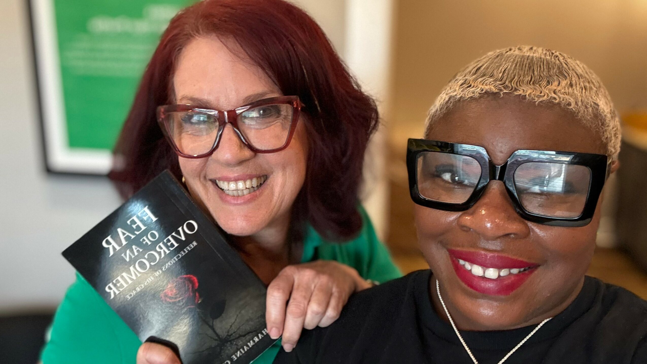 Two women smiling, one holding a book, both wearing glasses, indoors with a blurred background.