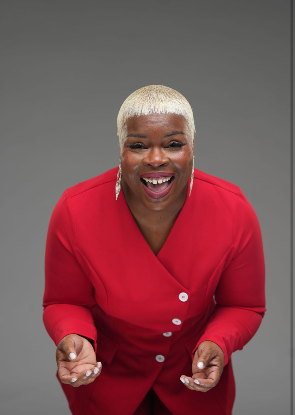 Smiling woman with short blonde hair wearing a red top, looking directly at the camera, gray background.