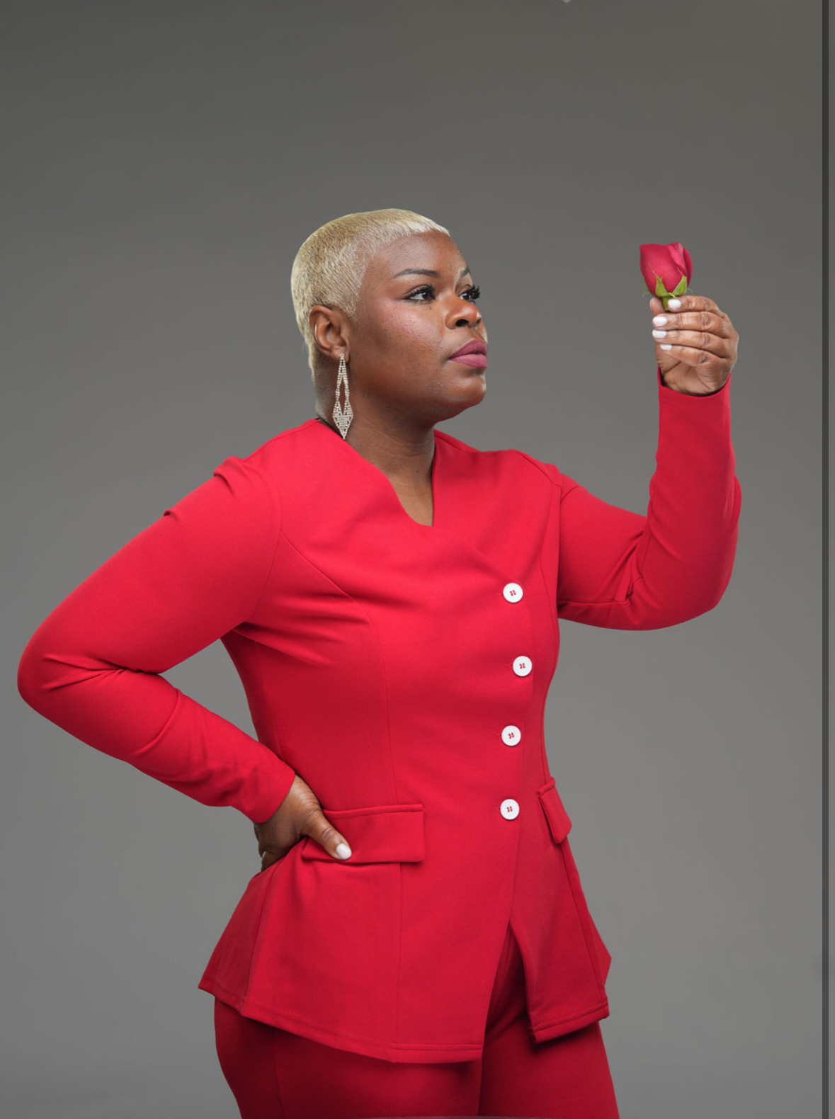 Woman with short blonde hair in a red blazer holding a small red flower, looking at it, against a gray background.