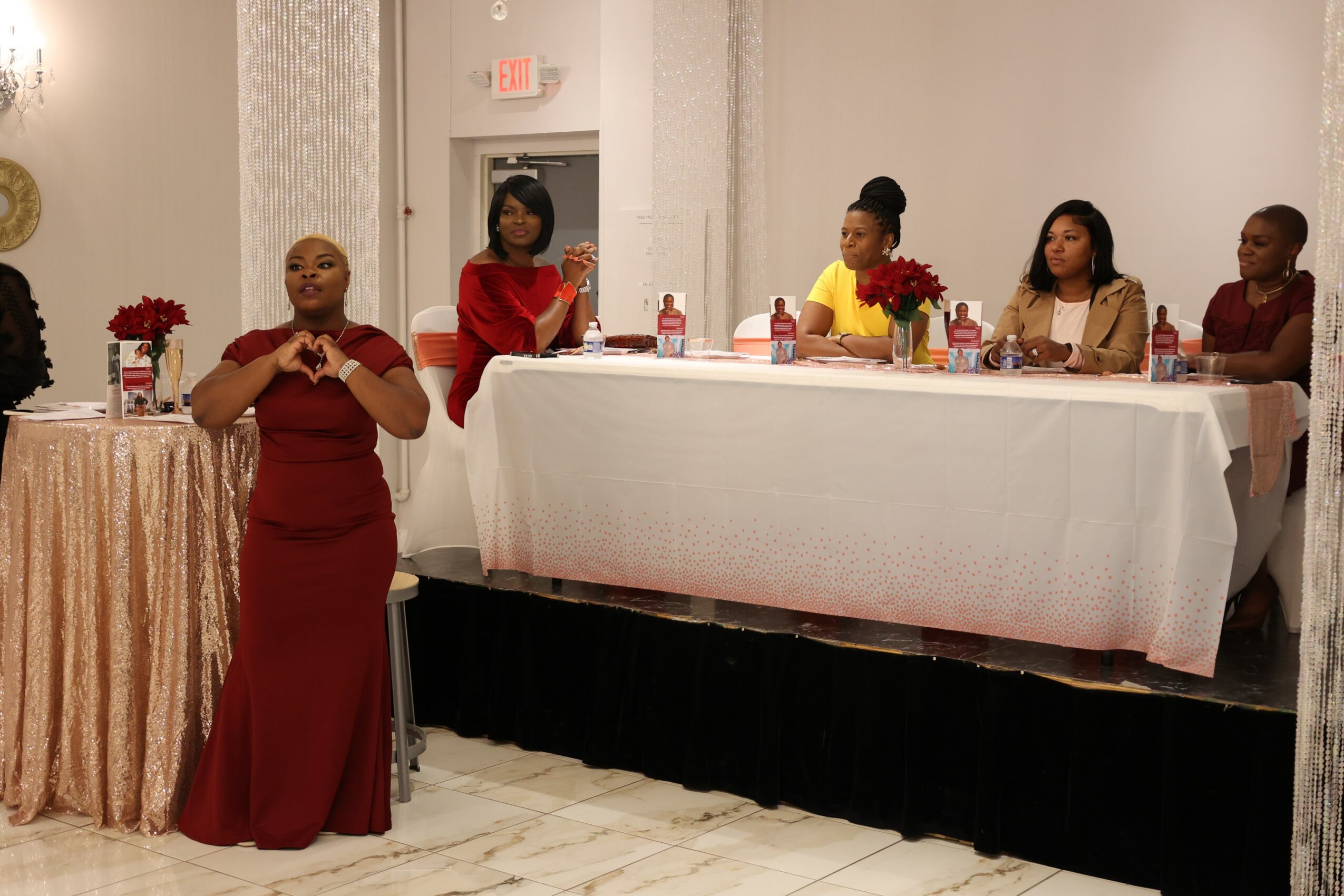 A woman in a red dress speaks at a formal event with a panel of five women seated at a table behind her, decorated with flowers.