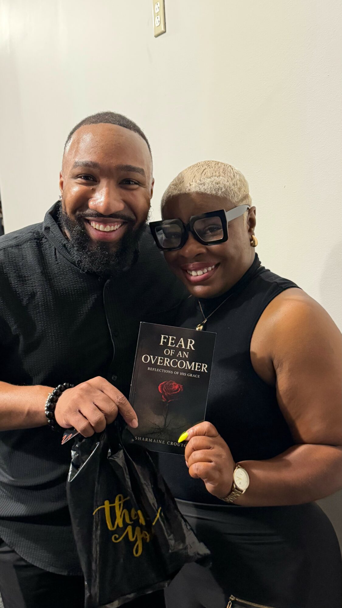 Two smiling people holding a book titled 'Fear of the Overcomer' and a black bag, standing against a plain wall.