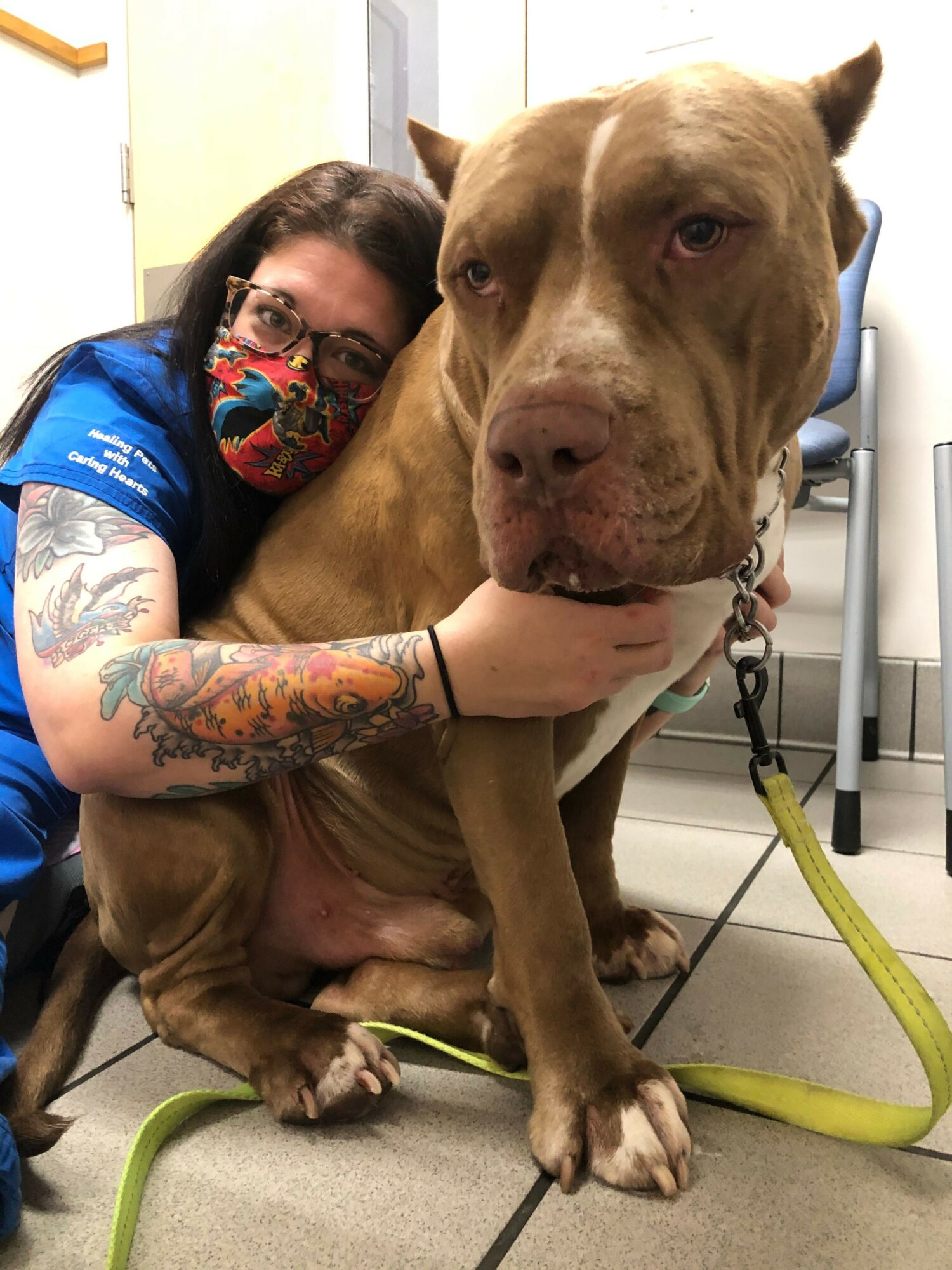 Person with tattoos and a colorful face mask hugging a large brown dog on a gray floor, with a yellow leash nearby.