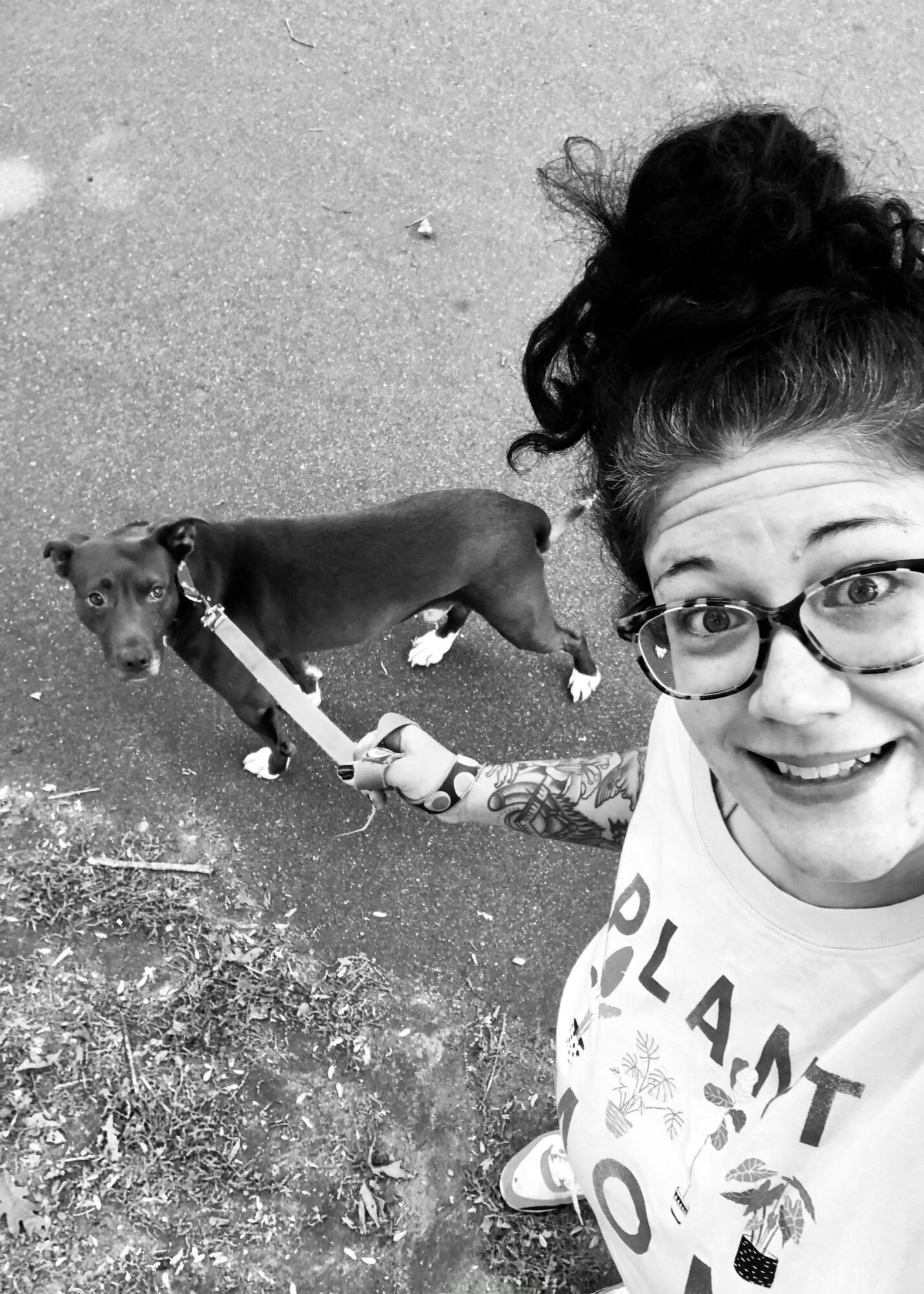 Woman with glasses taking a selfie outdoors, holding a leash attached to a small dog on a paved surface.