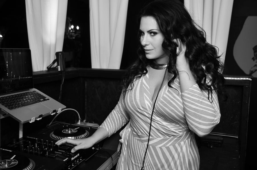 Woman with long curly hair wearing striped shirt, standing at DJ equipment in a room with curtains.