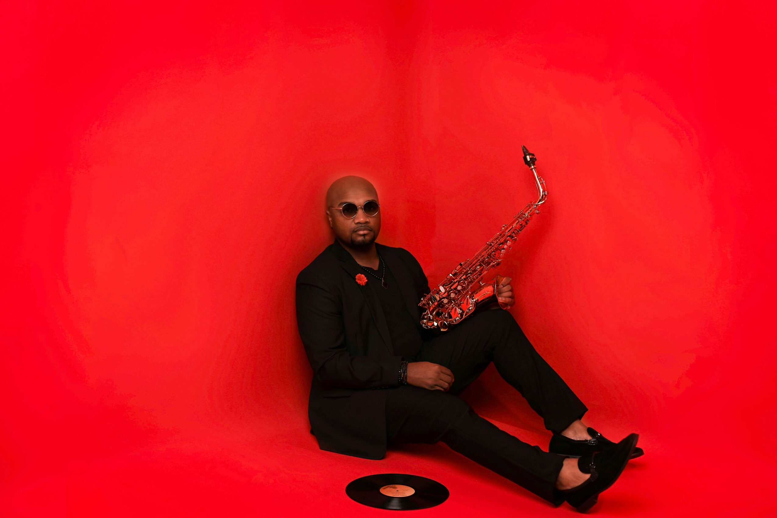 Person sitting on the floor holding a saxophone against a red background, wearing sunglasses and black clothing.