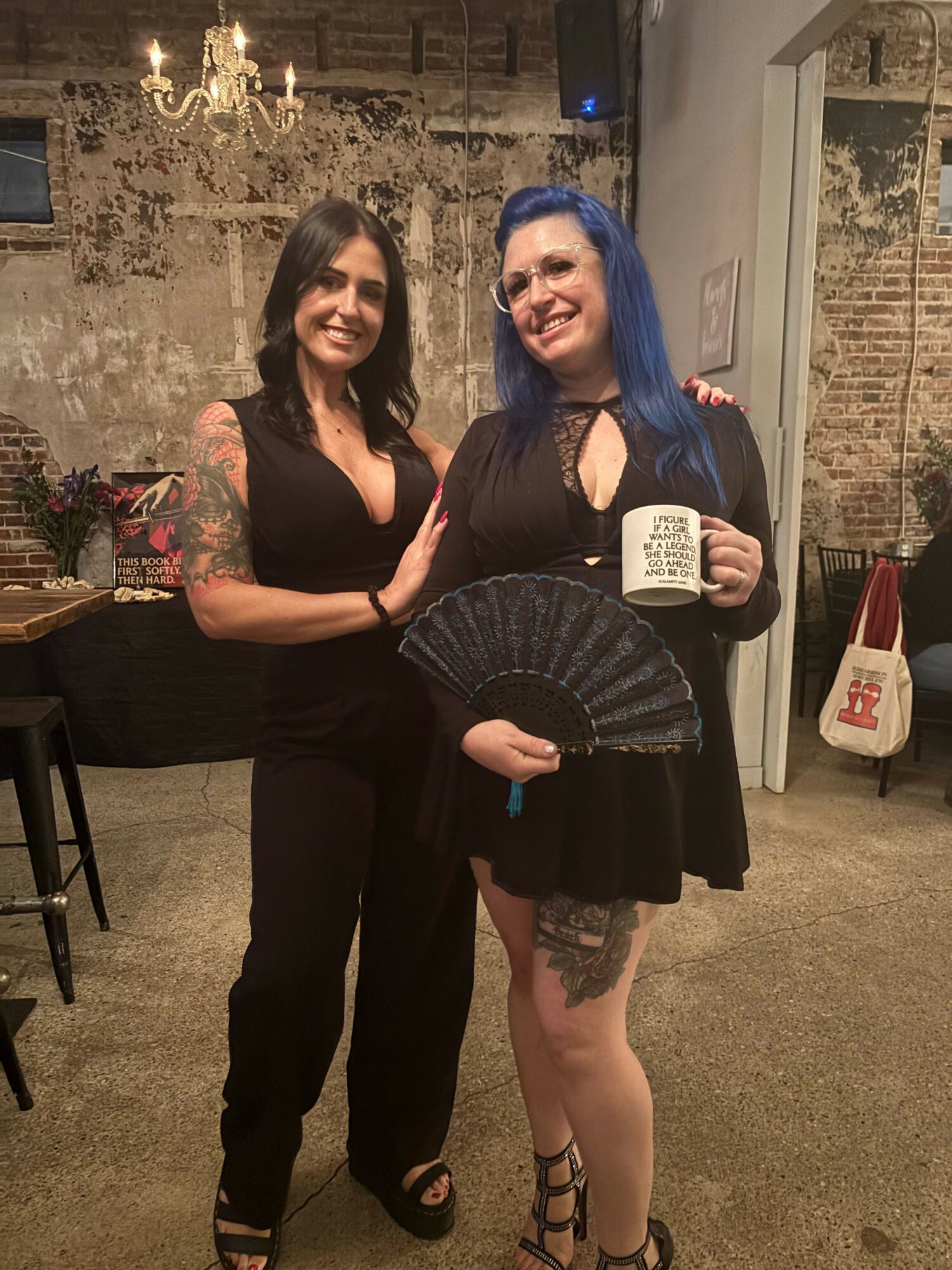 Two women standing indoors, one holding a fan and a notebook, the other with tattoos, both smiling.