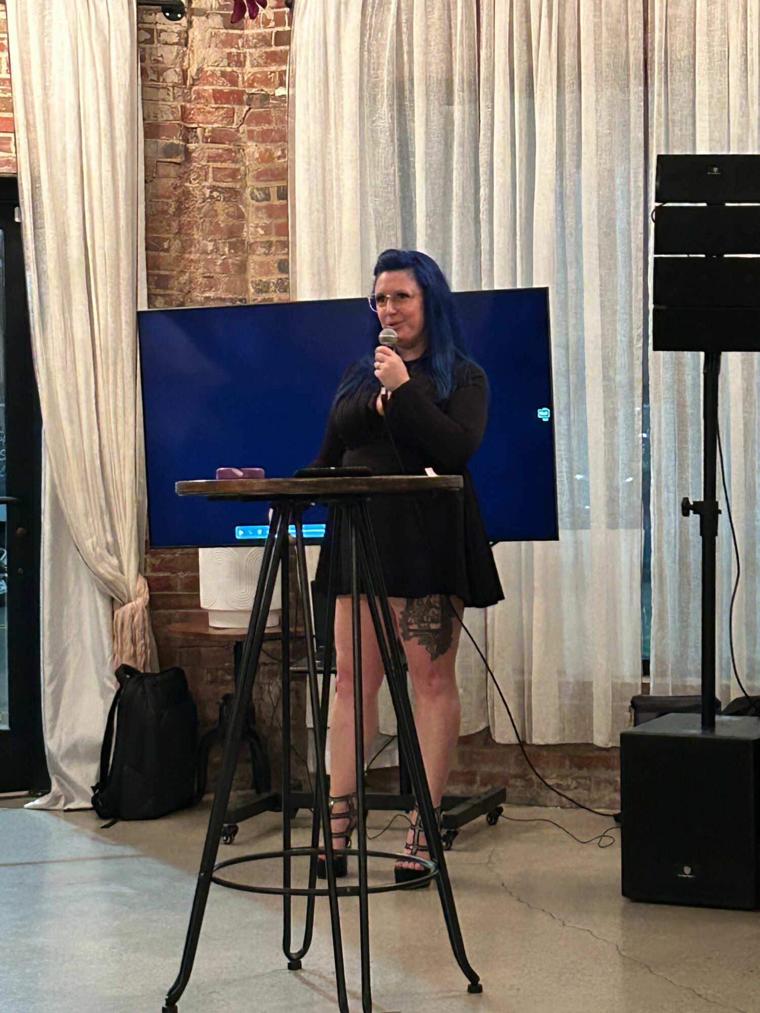 Woman standing at a microphone, speaking or singing, with a large screen behind her, in a room with brick wall and curtains.