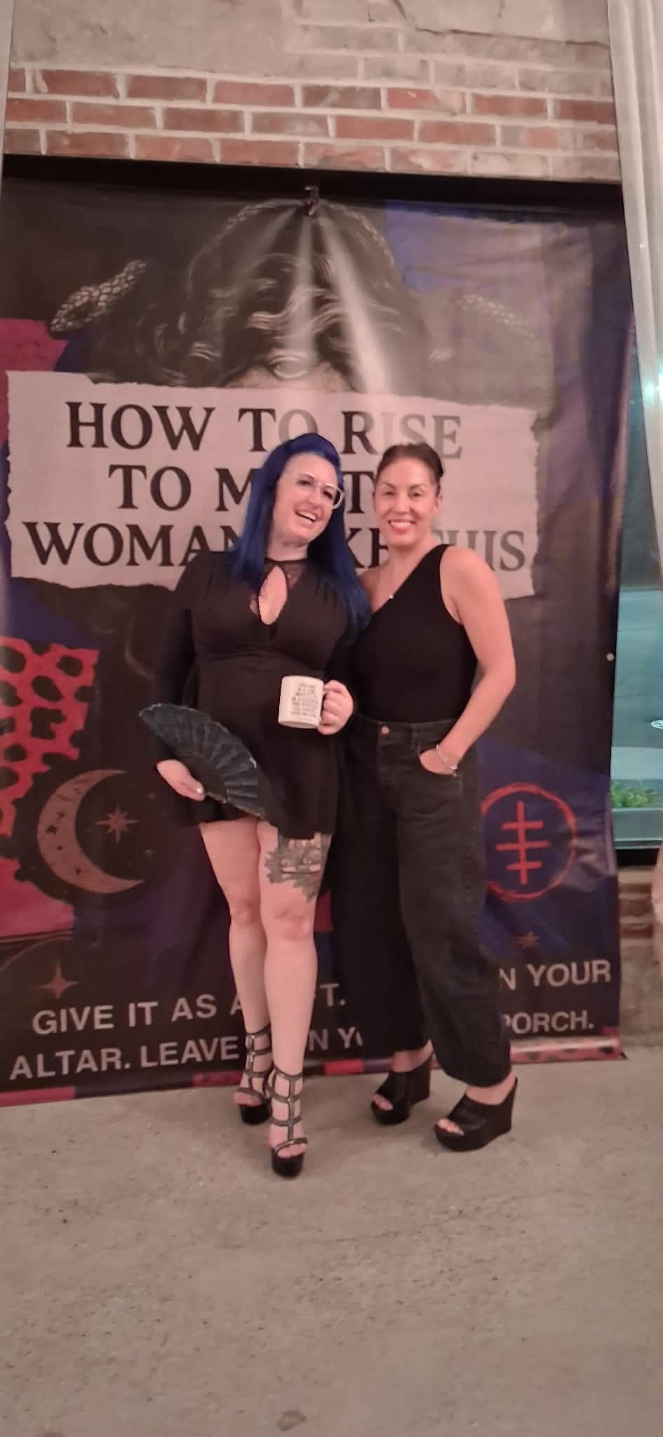 Two women standing in front of a banner, smiling, one holding a mug, both dressed in black, brick wall background.