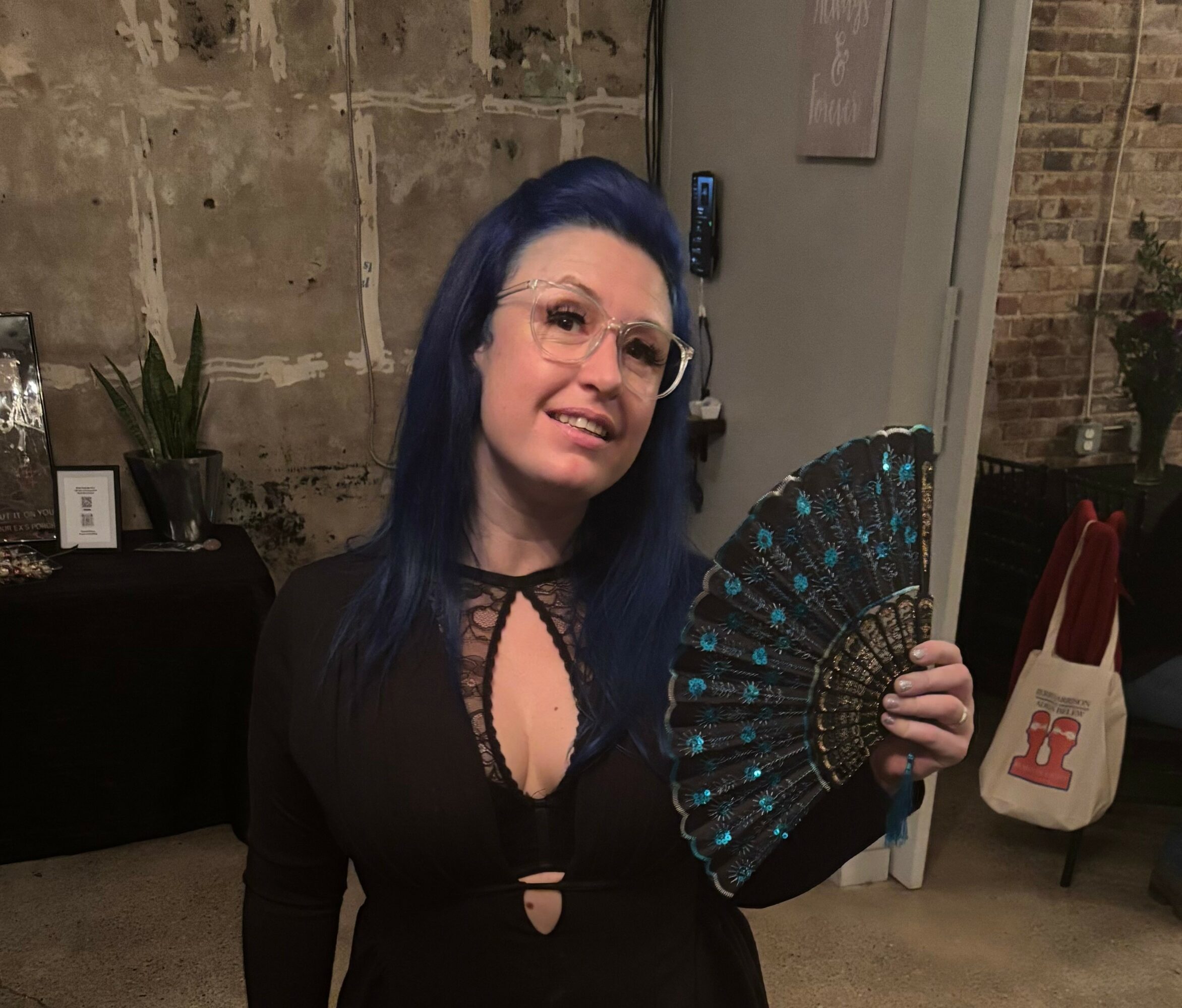 Woman with dark hair and glasses holding a decorative fan, standing indoors against a brick wall.