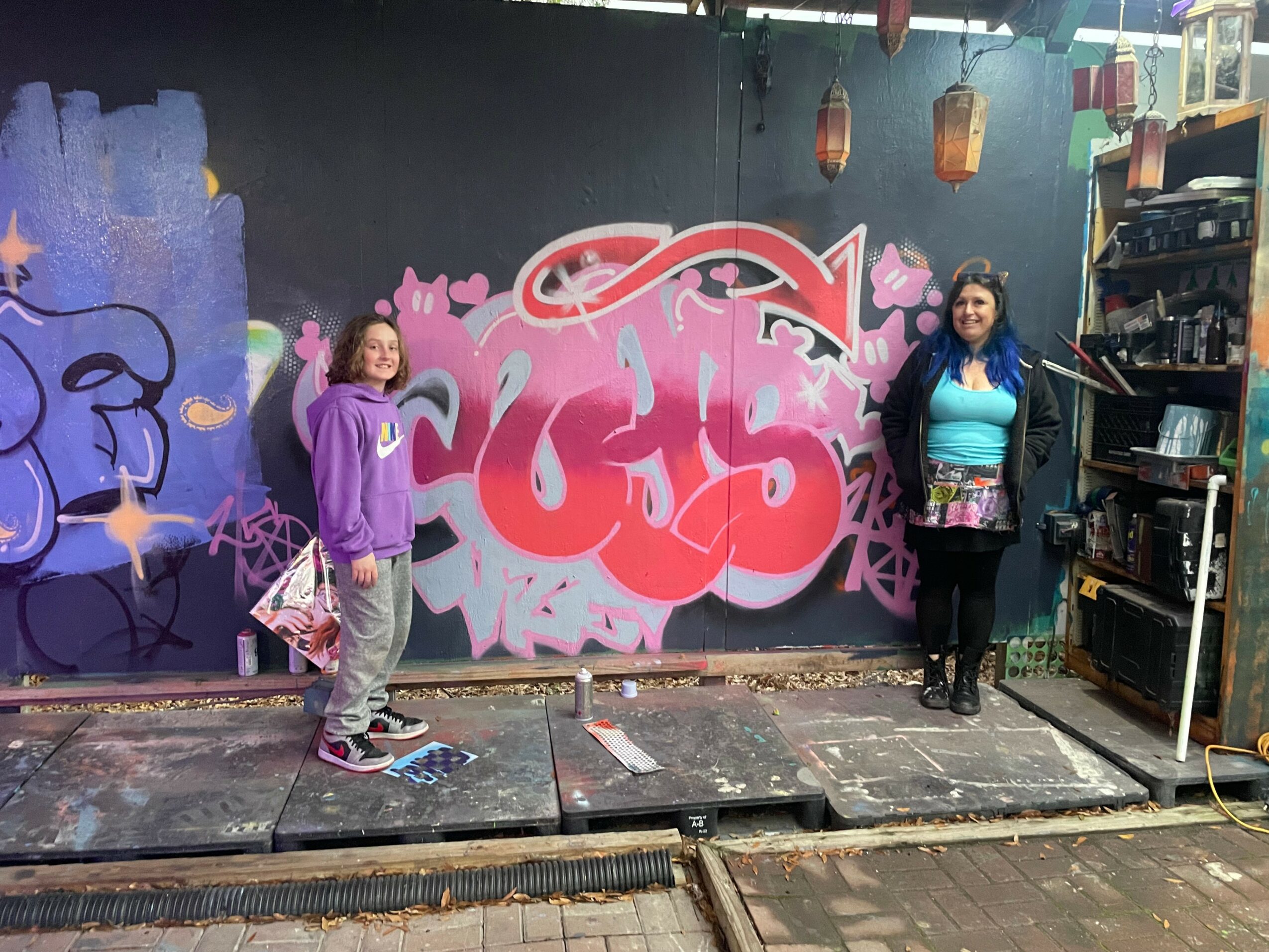 Two girls stand in front of a graffiti wall with pink and purple designs, one wearing a purple hoodie and the other a blue jacket.
