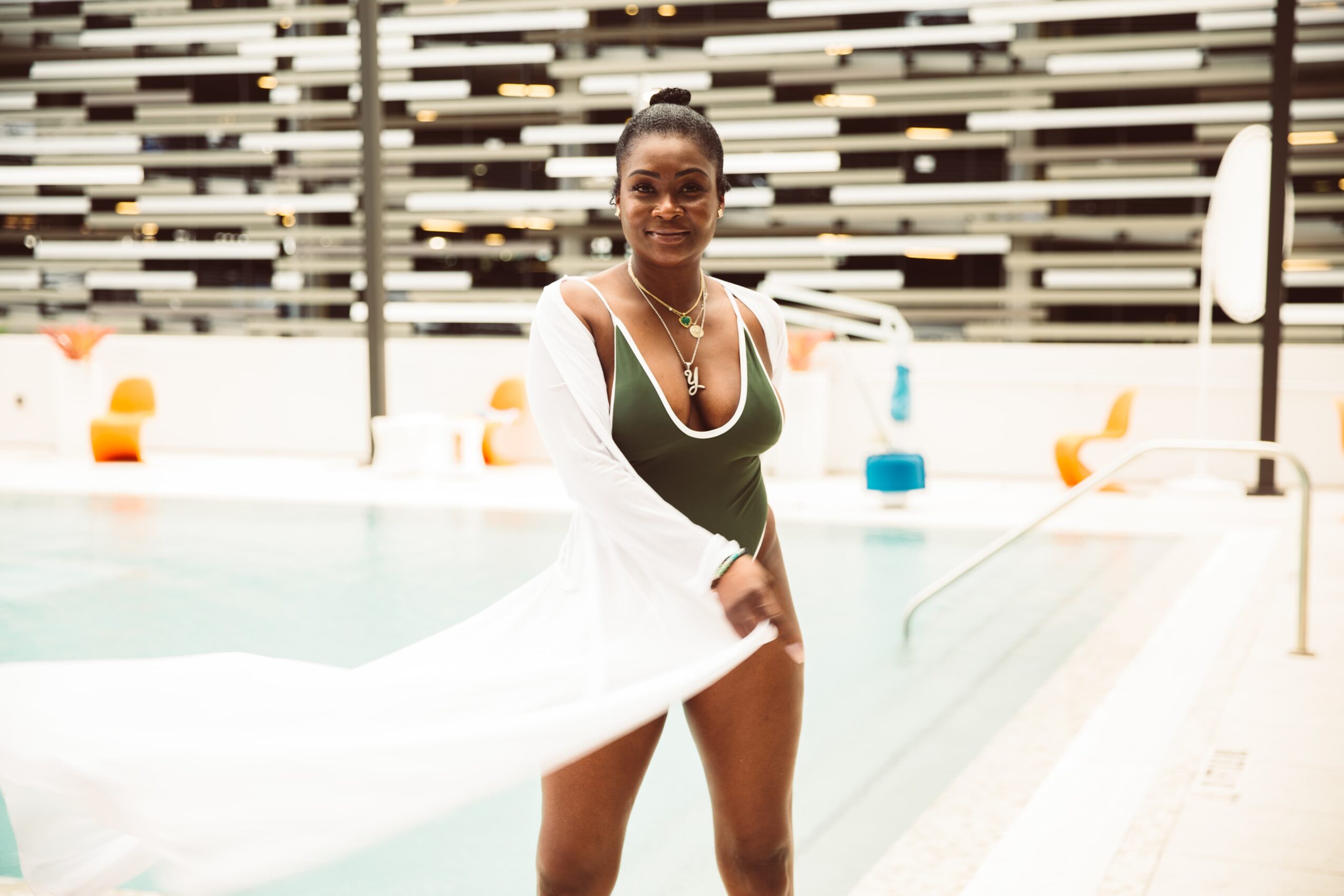 Woman in swimsuit standing by poolside with a white cover-up, smiling, with pool and modern background behind her.