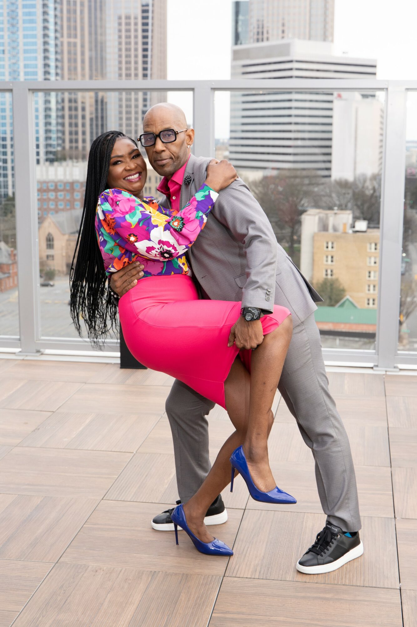 A man and woman dancing on a balcony with city buildings in the background, woman in a colorful top and pink skirt, man in gray suit.
