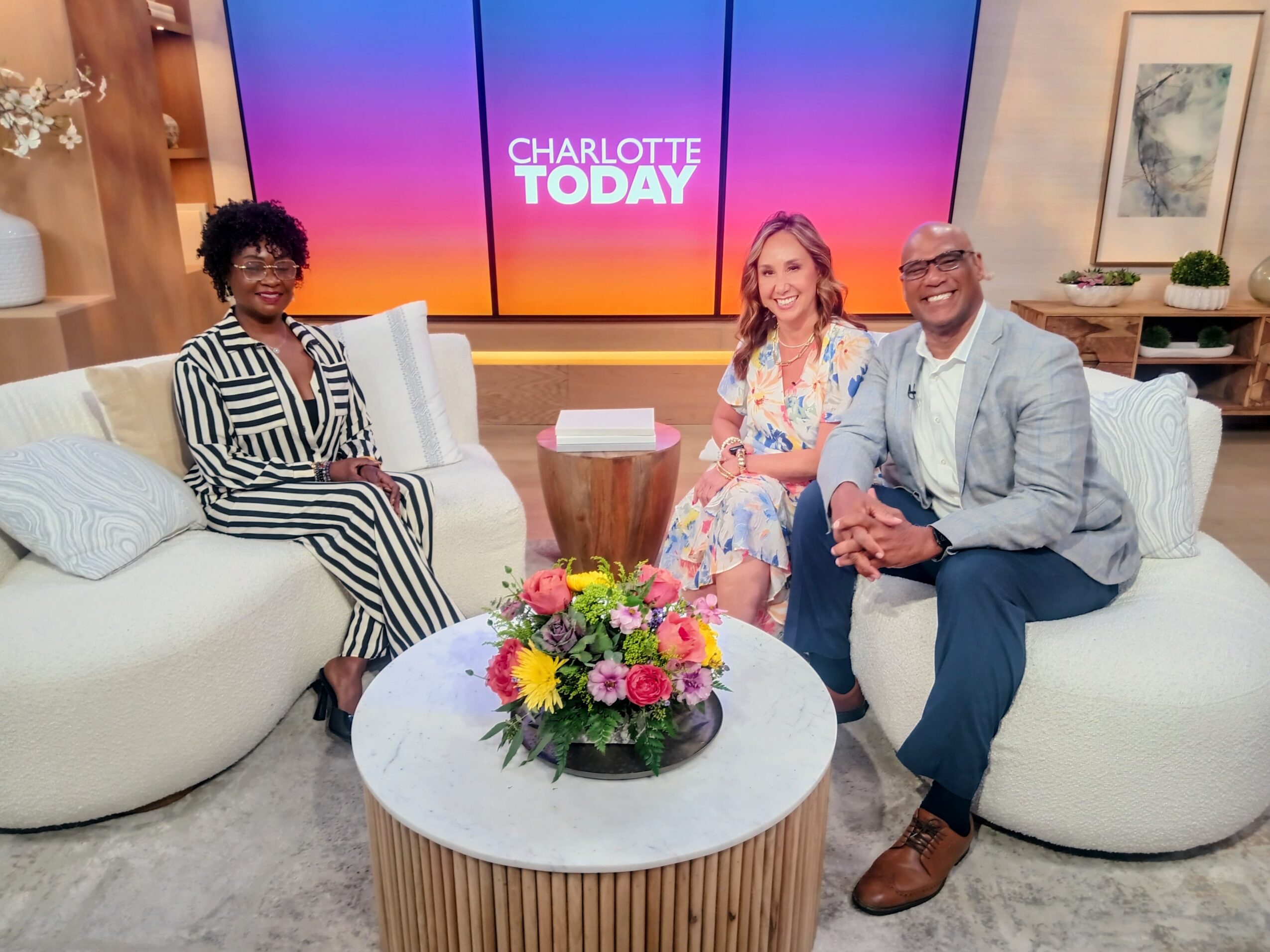 Three people sitting on a white sofa with a colorful flower arrangement on a round table in front, TV screen behind displaying 'Charlotte Today'.