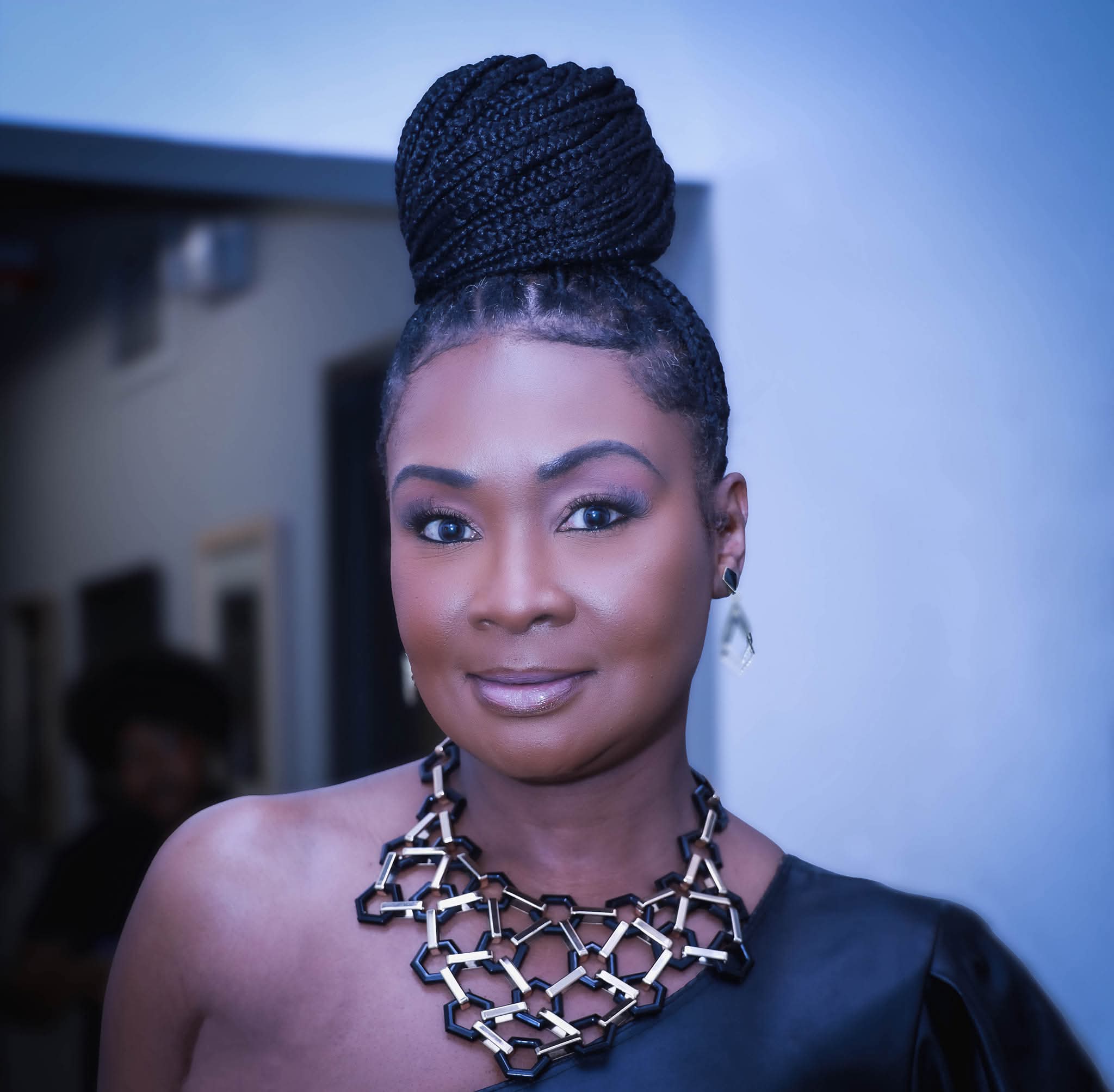Woman with braided hair styled in a high bun, wearing a statement necklace and a black off-shoulder top, smiling softly.
