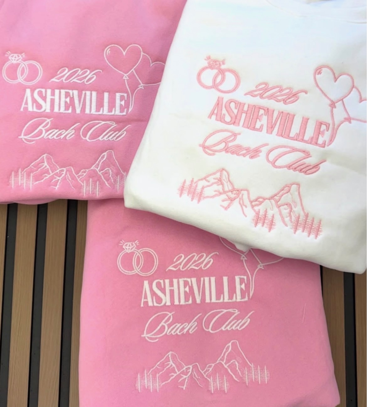 Three folded shirts with Asheville Bach Club embroidery, pink and white, on a wooden surface.