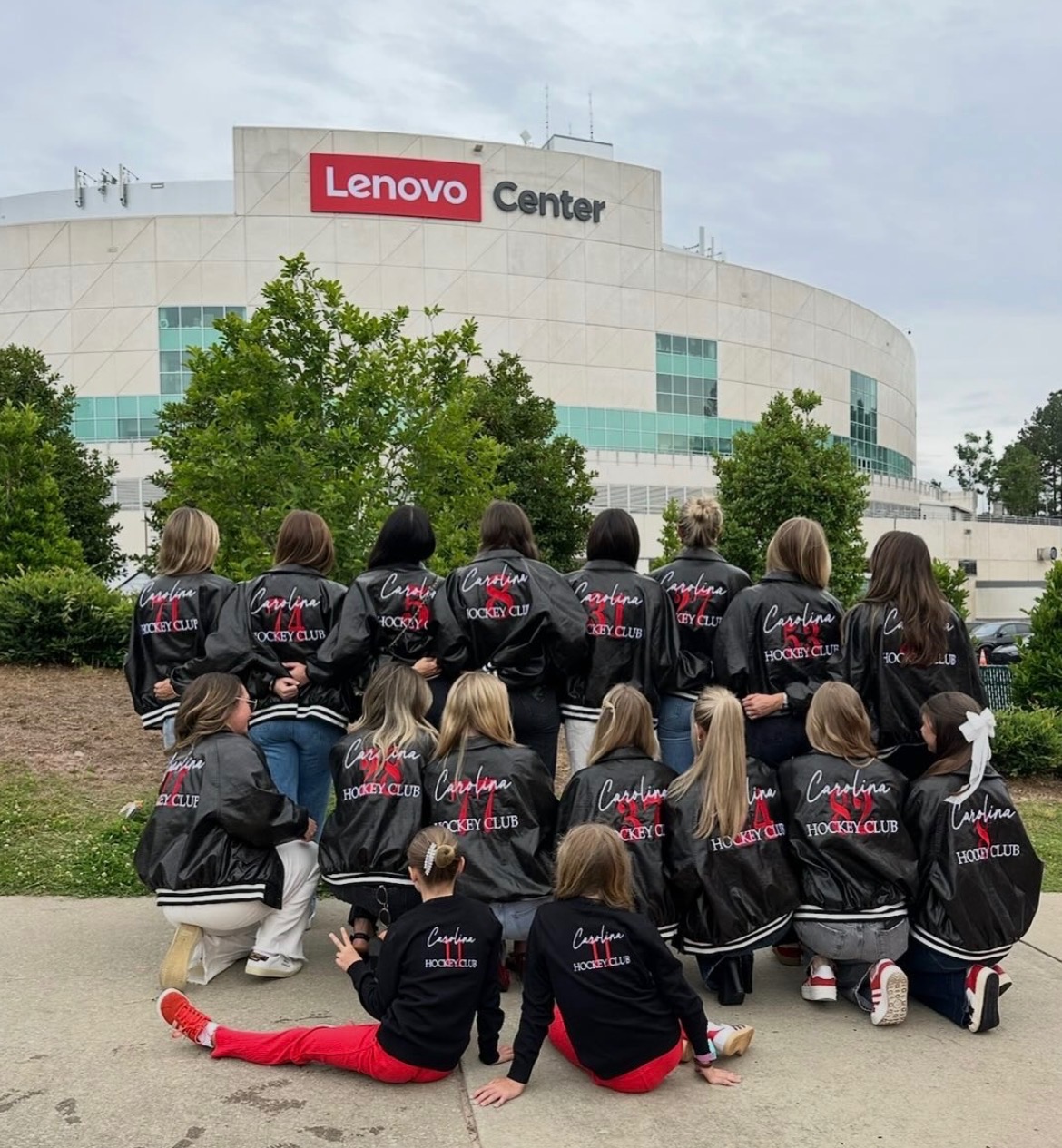 Group of people wearing matching jackets in front of Lenovo Center building with trees and cloudy sky