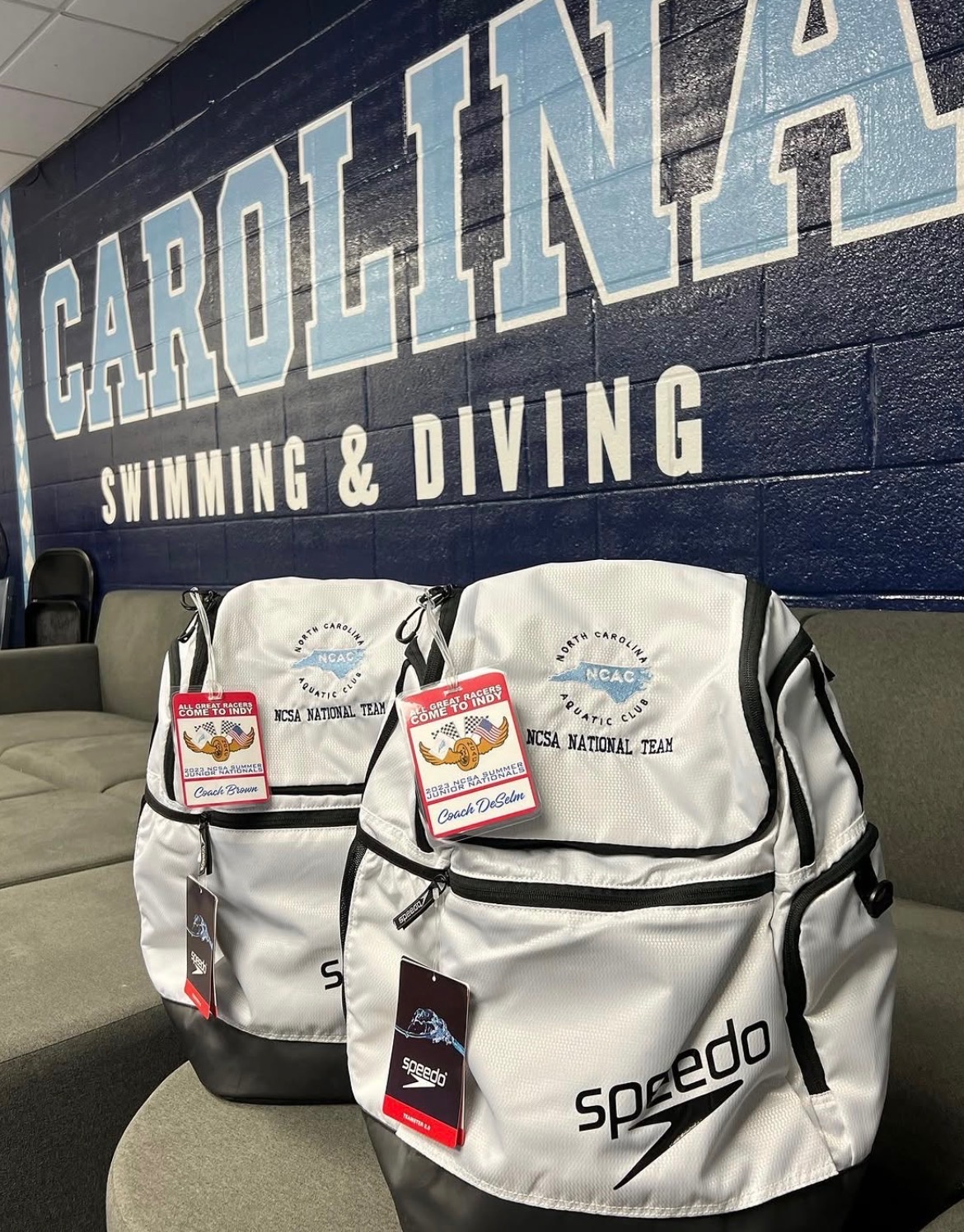 Two white Speedo backpacks with tags, placed on a black bench against a blue wall with white text reading 'CAROLINA SWIMMING & DIVING'.