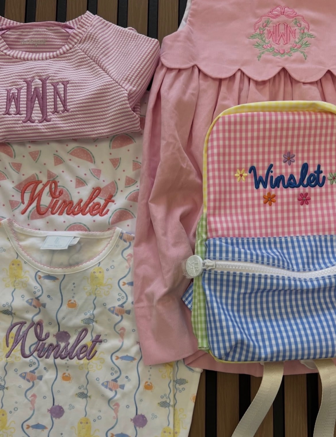 Four children's clothing items and a backpack on a striped surface, including pink, white, and patterned fabrics.