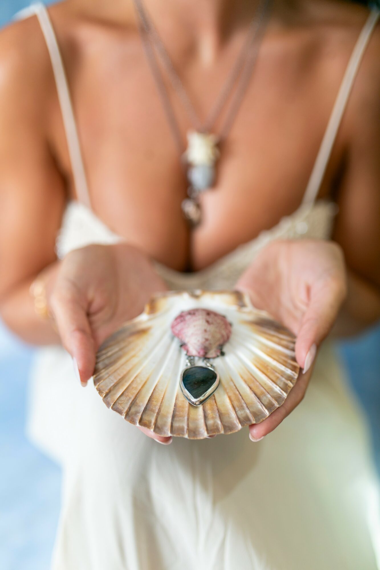 Person holding a seashell with a heart-shaped object inside, wearing a white dress and layered necklaces.