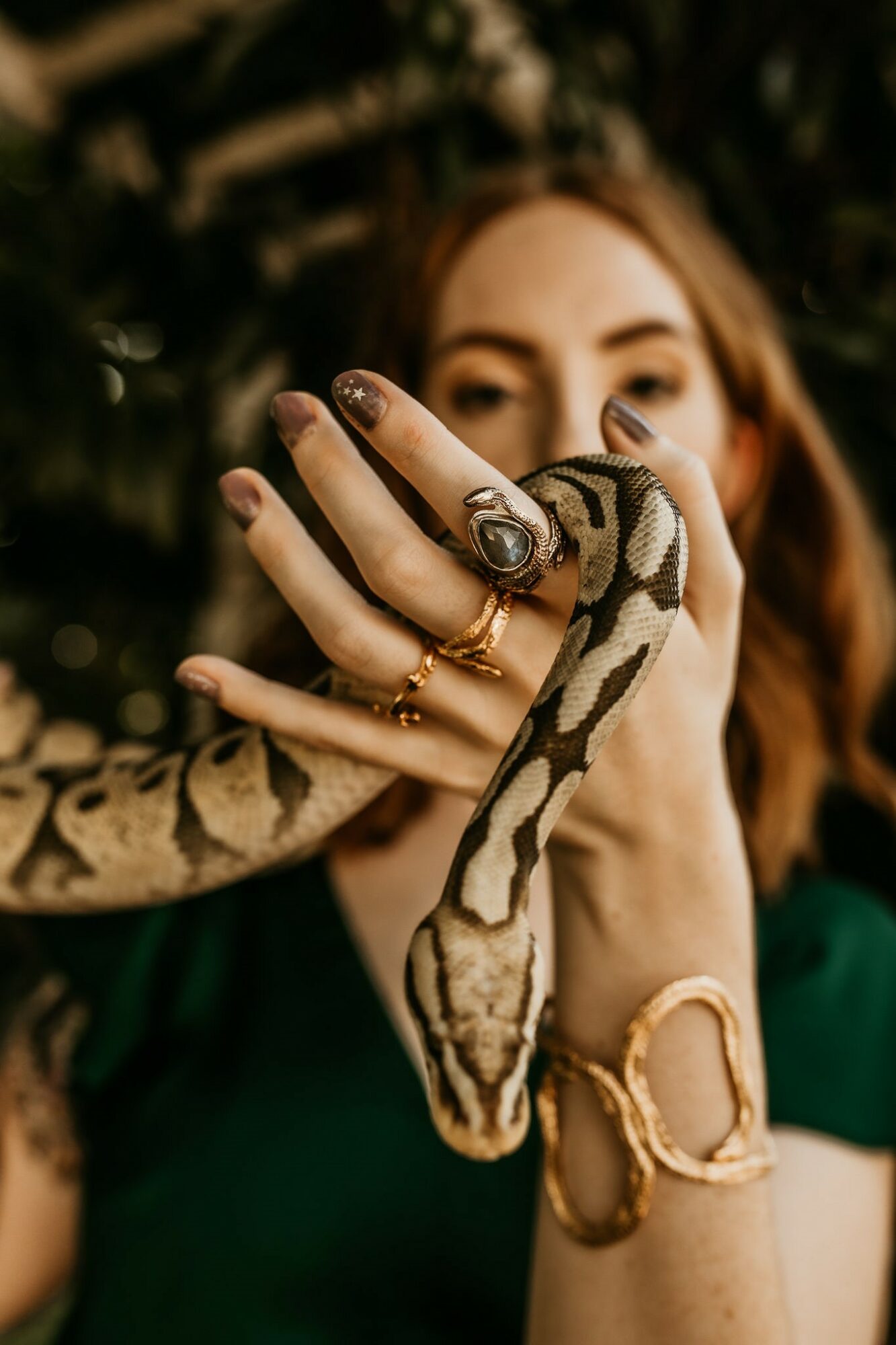 Person holding a snake with rings on fingers, outdoors with blurred greenery background.