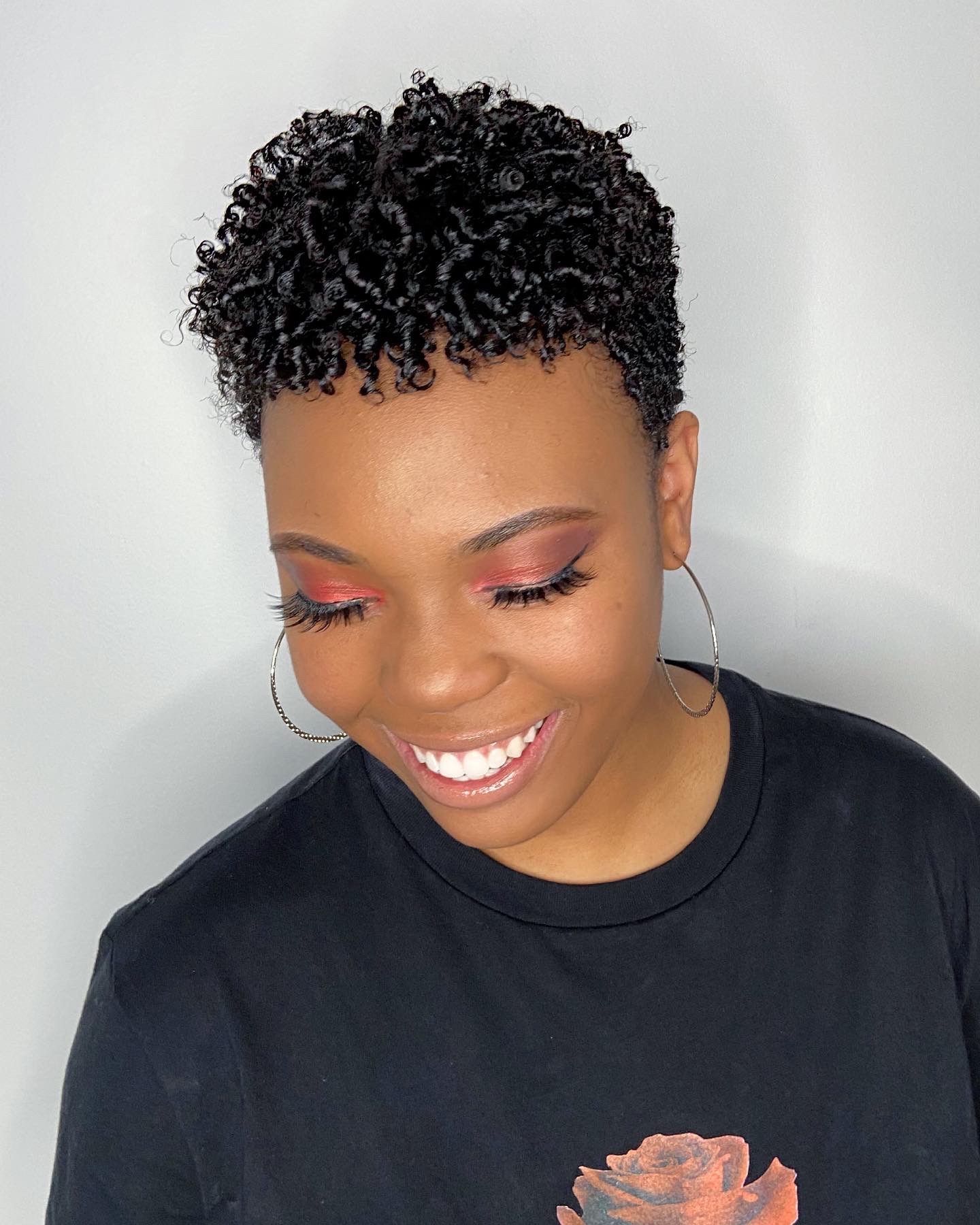 Smiling woman with short curly hair, wearing hoop earrings and a black shirt, looking down.