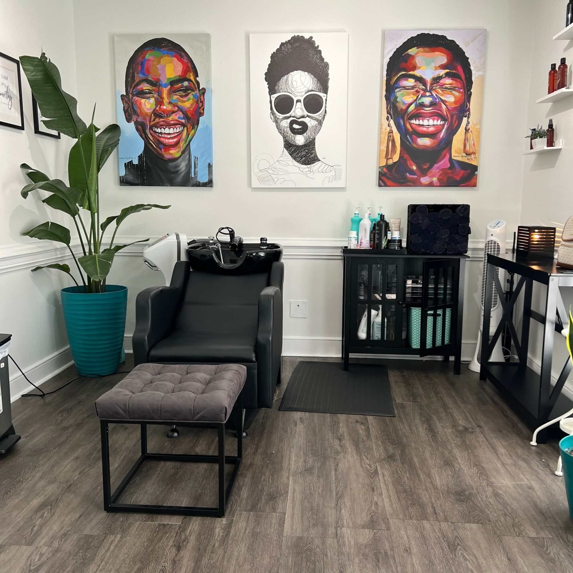 Interior of a salon with colorful portraits on the wall, a black chair, a gray ottoman, and a plant in a blue pot.