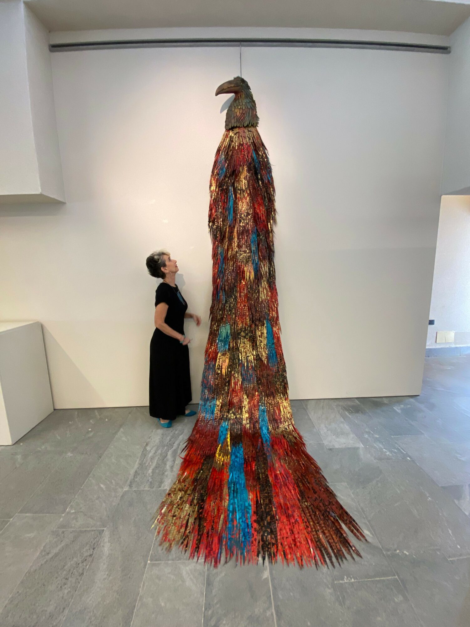 Person standing next to a colorful, textured sculpture resembling a peacock with a long tail, in an art gallery.