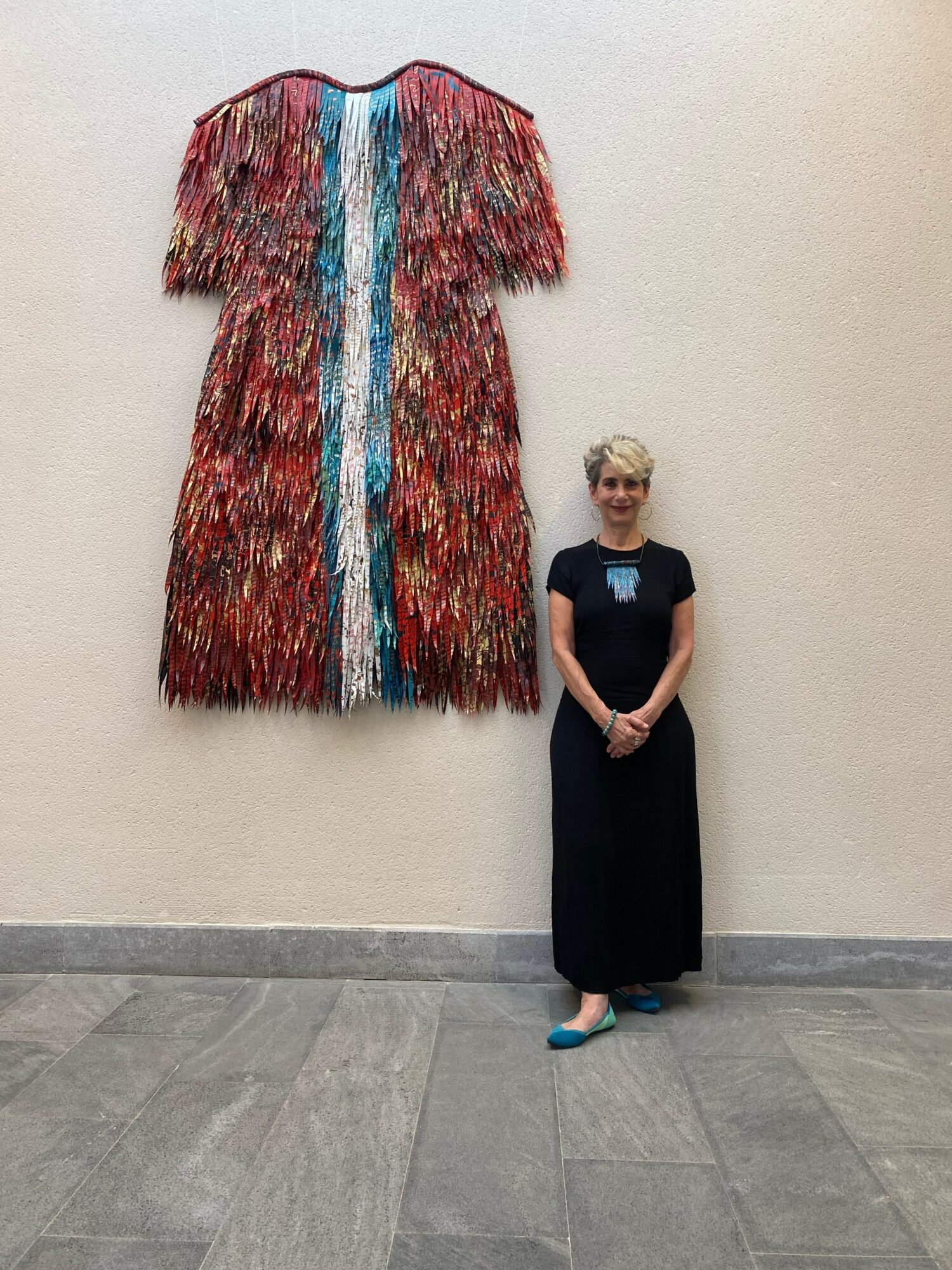 Person standing next to a colorful, textured coat or garment hanging on a wall, in an indoor setting.
