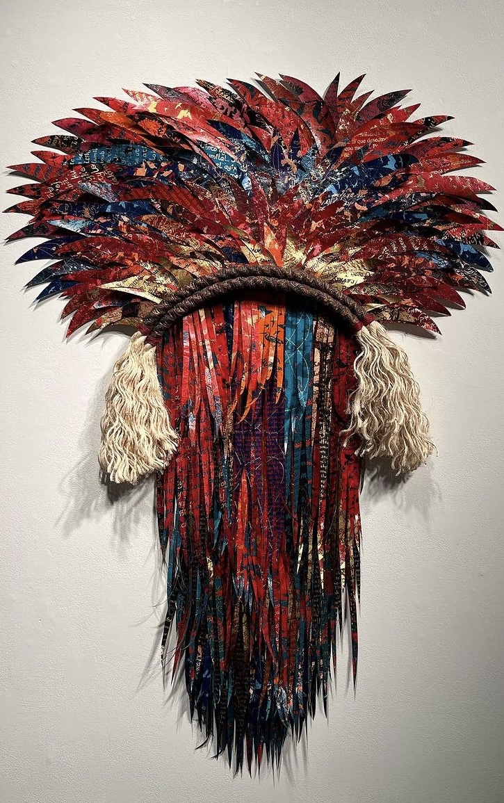 Colorful feathered headdress with hanging fabric and beige tassels, displayed on a white wall.