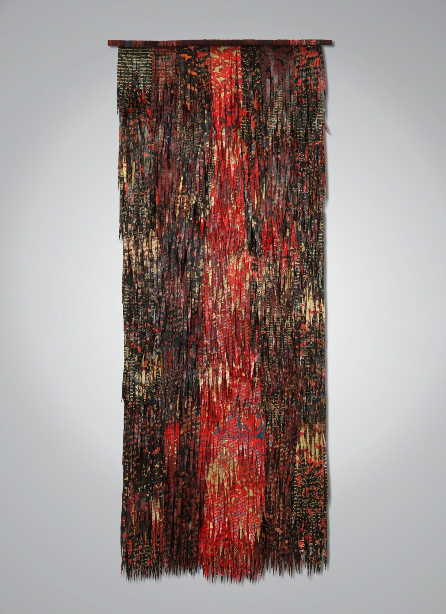 Long, vertical, multicolored woven textile with red, black, and gold hues, hanging from a rod against a plain background.