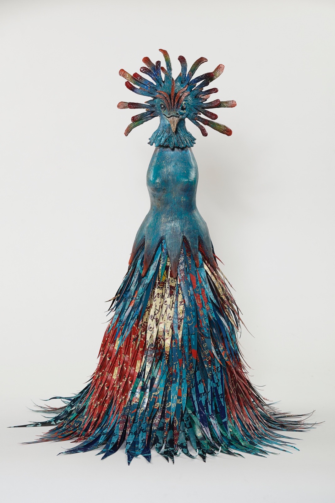 Colorful sculpture of a figure with a headdress and a long, feathered skirt, standing against a plain background.