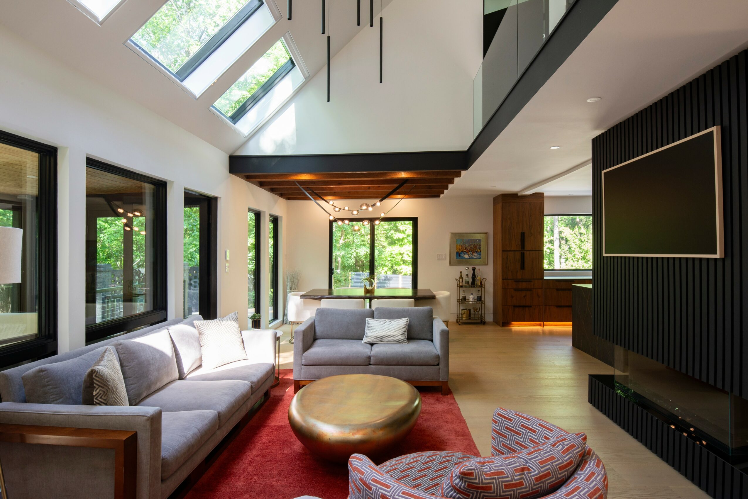 Living room with large windows, sofas, a round coffee table, and a flat-screen TV on a black wall panel.