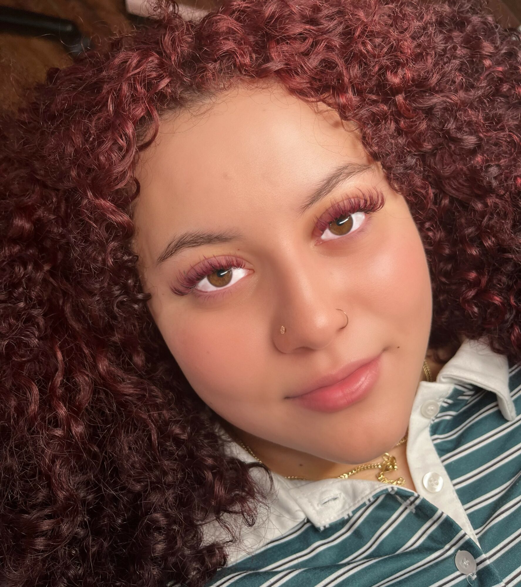 Close-up of a young woman with curly hair, wearing a striped shirt and gold jewelry, looking at the camera.