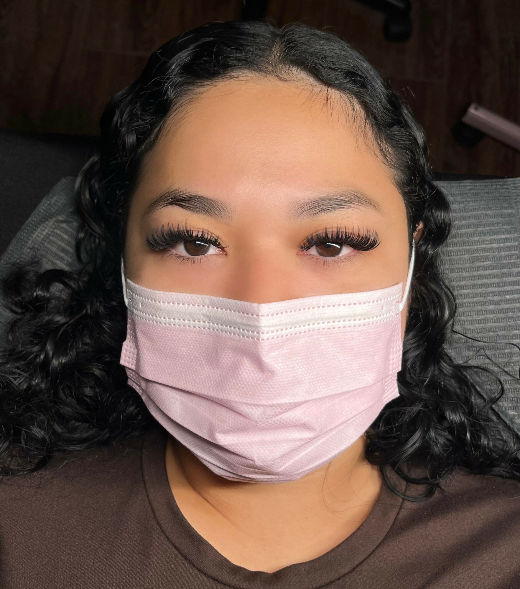 Close-up of a woman with dark curly hair wearing a pink face mask and brown shirt, looking directly at the camera.