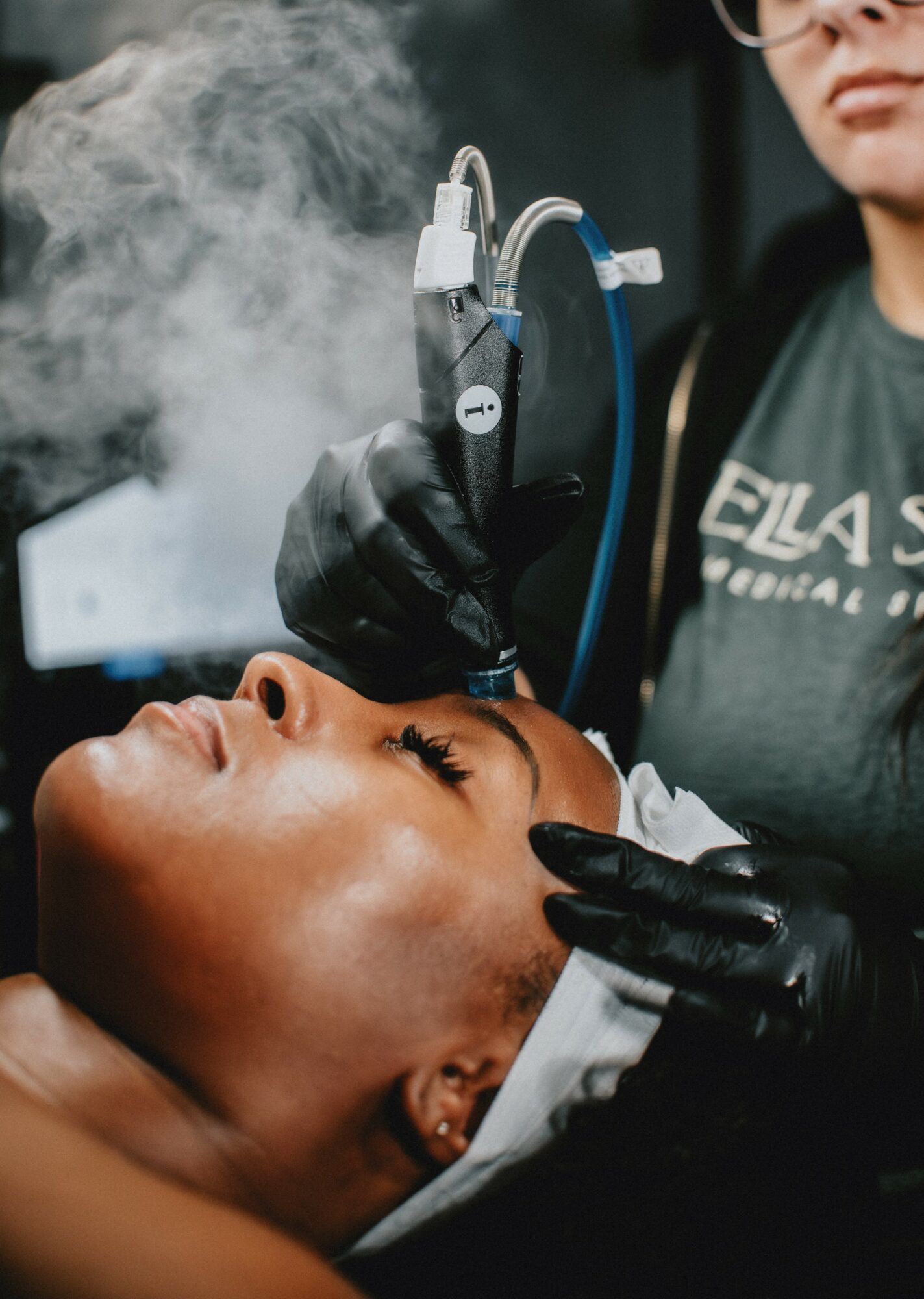 A person receives a cosmetic procedure with a device emitting vapor, while a technician in gloves operates the equipment.